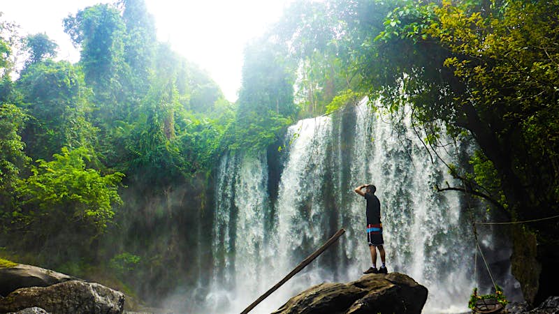 trekking-cambodia A man in shorts stands on a large rock in a forest; behind him is a huge waterfall .