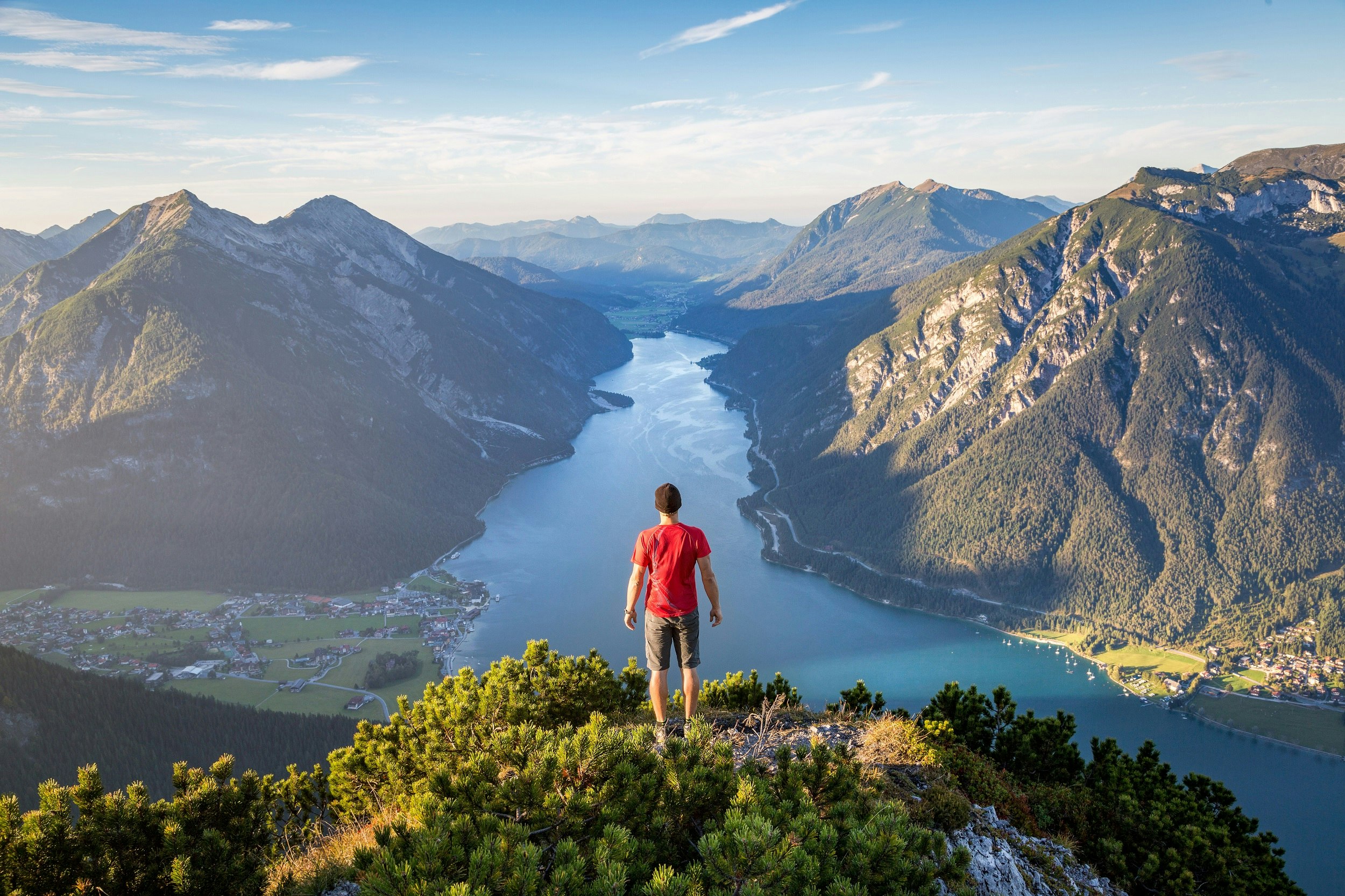 A man wearing shorts, t-shirt and black beany stands on an outcrop and looks out over a spectacular valley lake; mountains climb on all sides.