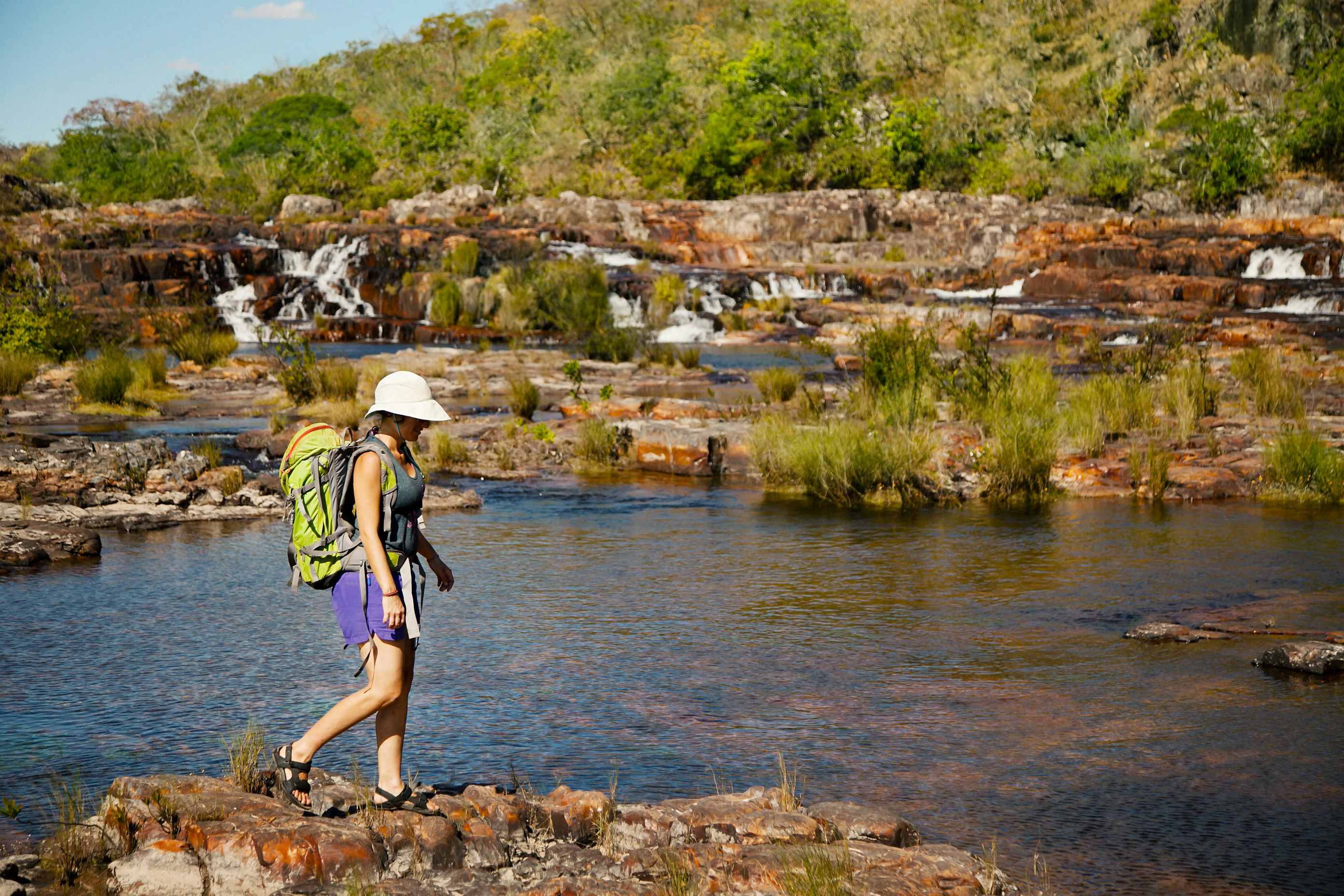 A woman hiking over rocks with a waterfall in the background at Chapada dos Veadeiros National Park.