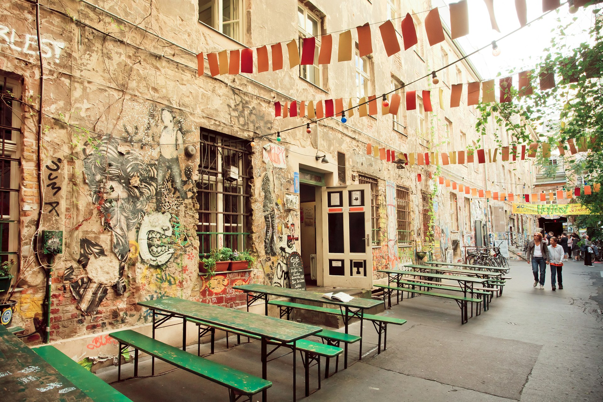 The graffiti-painted exterior of a trendy bar in Berlin. There are simple green tables and benches lined up outside and bunting is hanging up overhead.