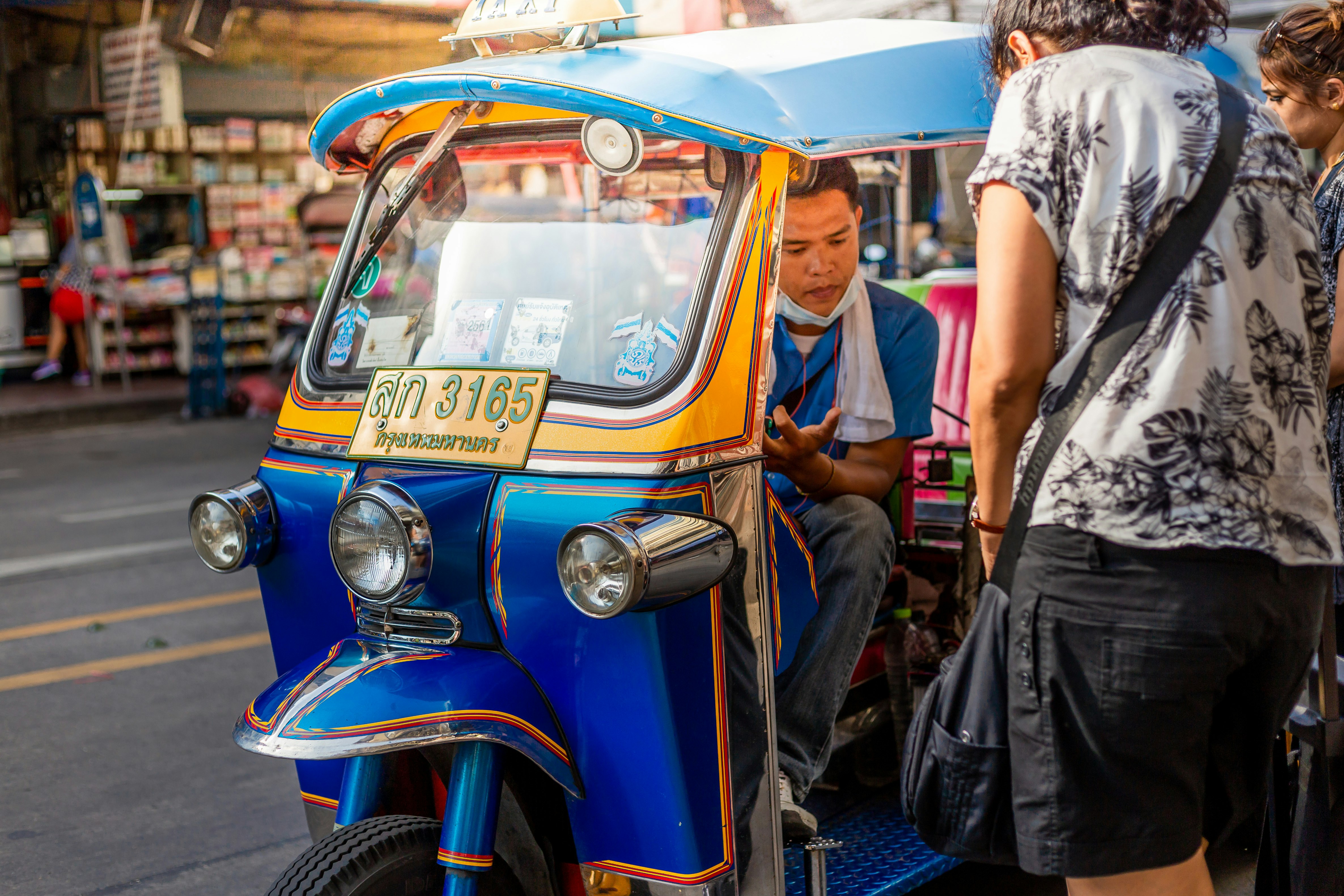 Two women consult with a tuk tuk driver at the side of a road in Bangkok. The small, striking vehicle is painted yellow and blue.
