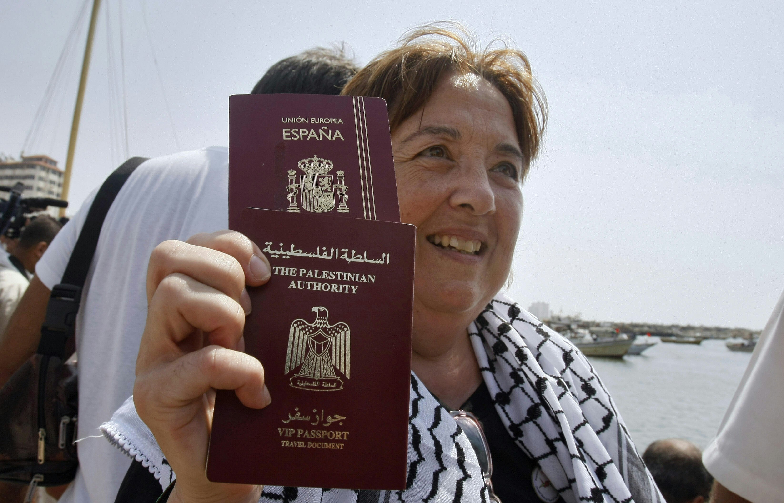 A woman with short light brown hair hold up two maroon passports with gold lettering, one for Espana and one from the Palestinian Authority. She is wearing a white and black scarf and is standing by the sea in Gaza