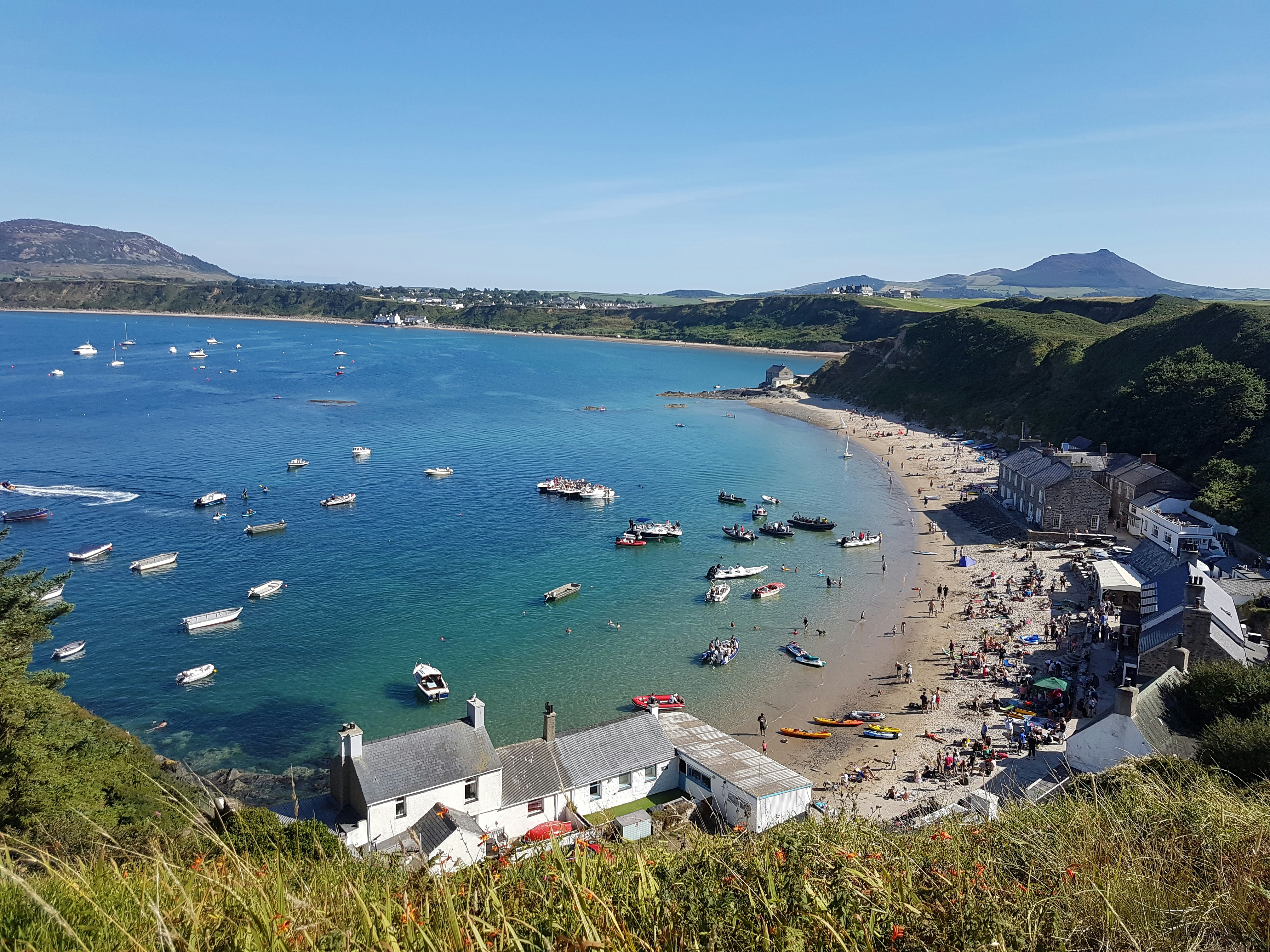 A view looking down to the boat-dotted bay at the busy Morfa Nefyn beach. Buildings line the beach-front, and many people are milling around on the sand outside Tŷ Coch Inn.