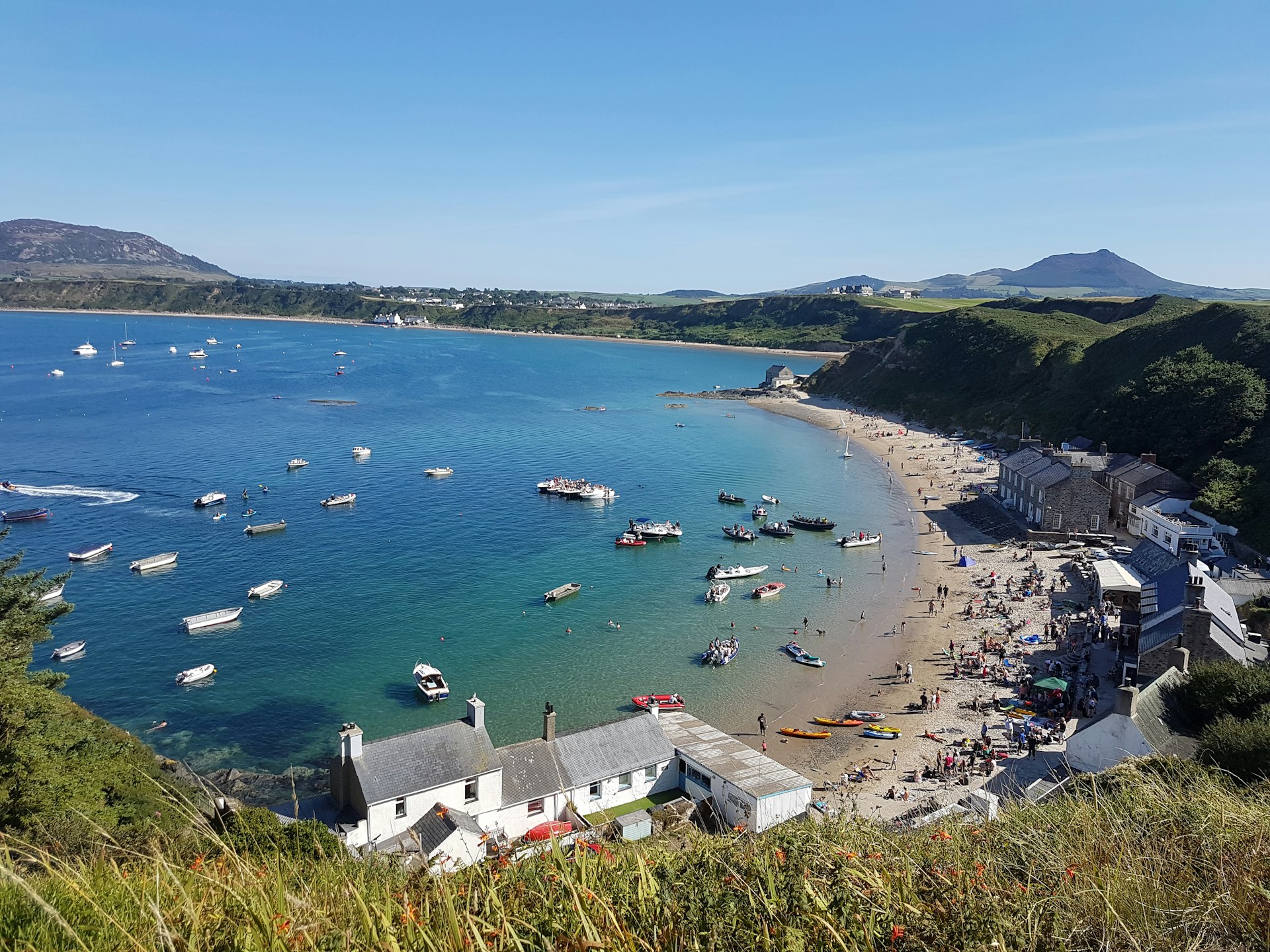 A view looking down to the boat-dotted bay at the busy Morfa Nefyn beach. Buildings line the beach-front, and many people are milling around on the sand outside Tŷ Coch Inn.