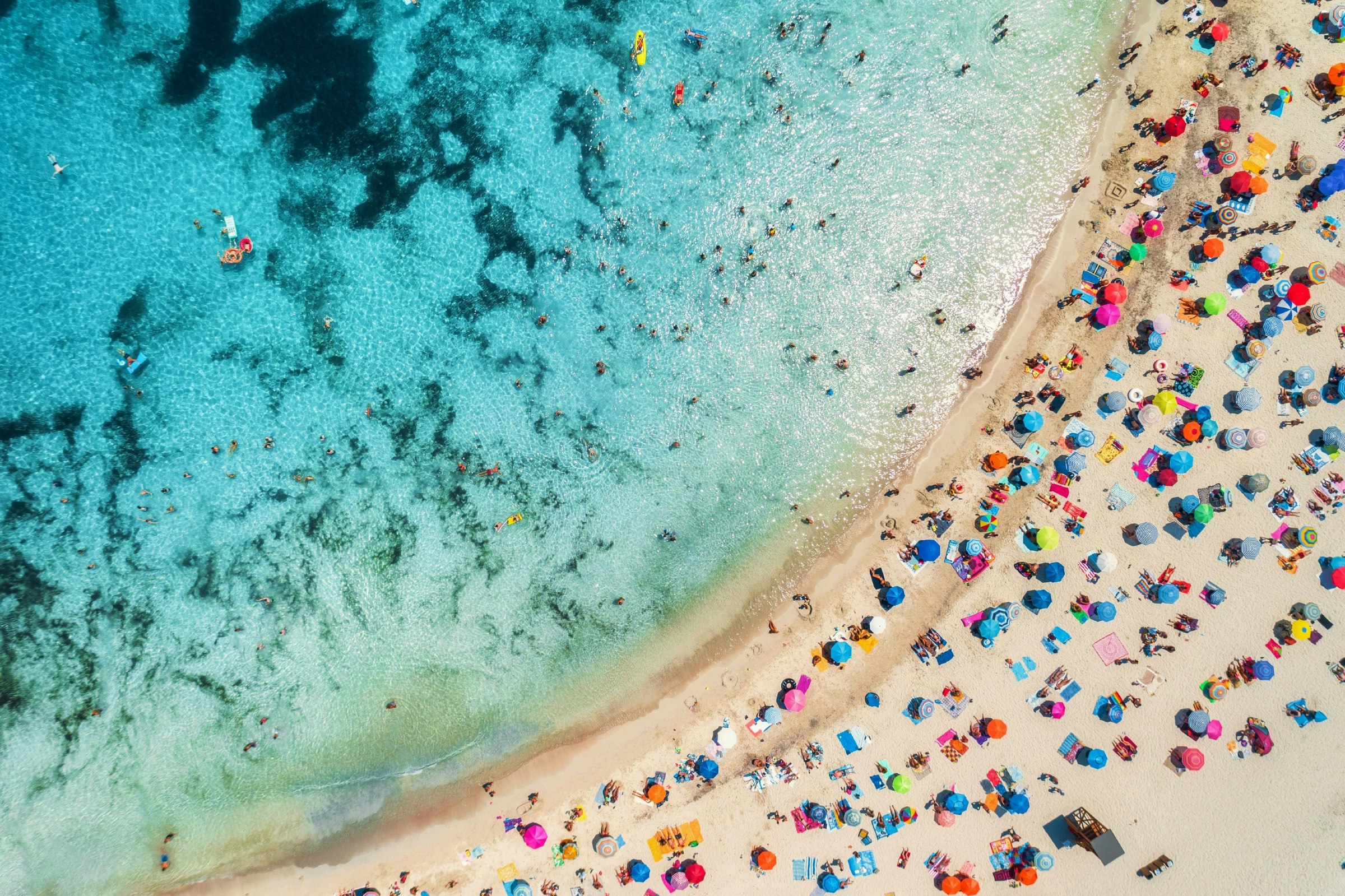 Aerial of a crowded sandy beach with colourful umbrellas, sun bathers and swimmers during summer.