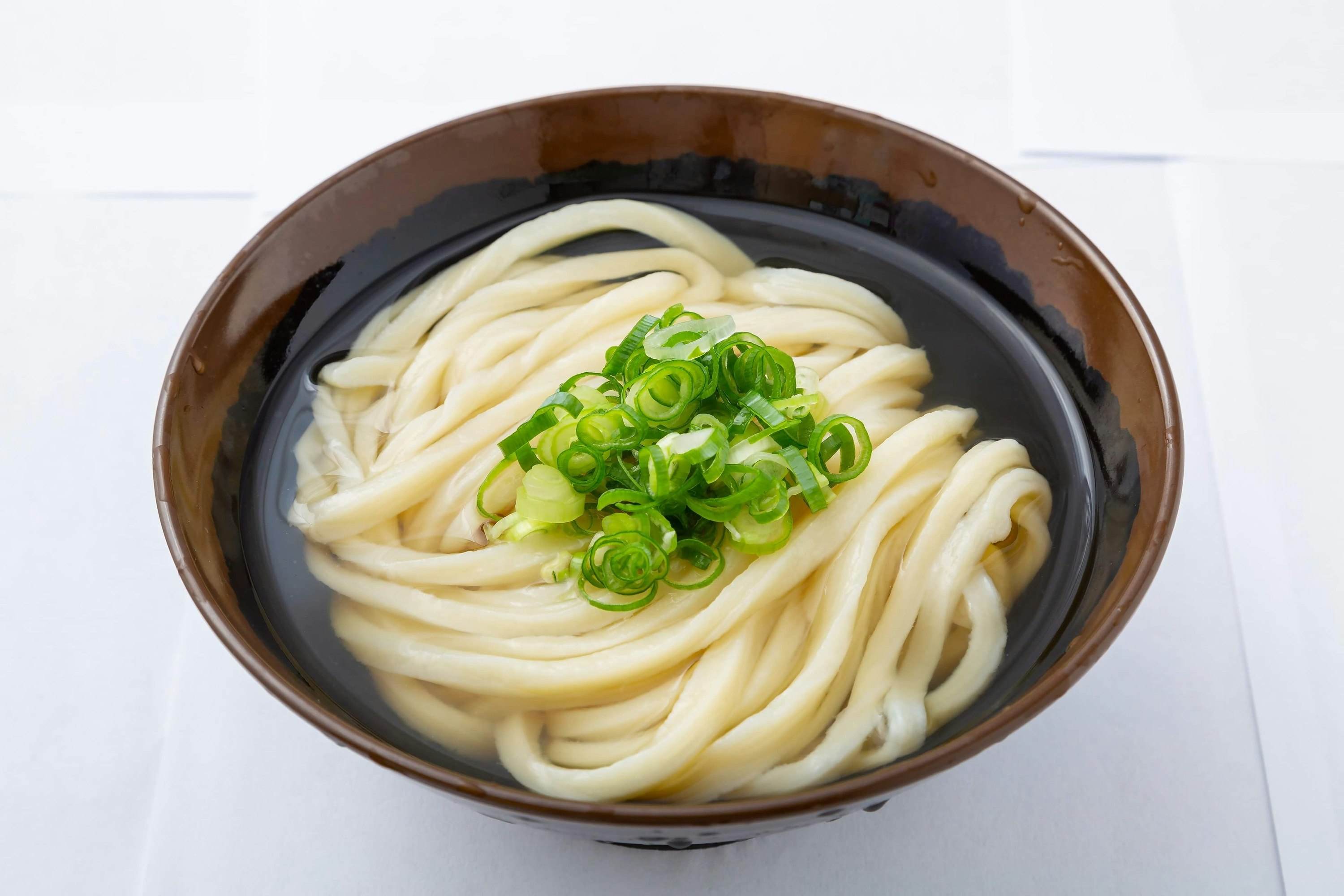 A grown and navy blue glazed bowl sits against a white backdrop and holds a clear broth and a tidy mound of pale udon noodles topped with a bright green cluster of shaved green onions