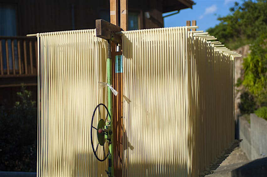 udon-noodles-shodoshima-somen-kagawa-japan.jpg Long strands of shodoshima somen noodles hang from racks outdoors to dry in the sun. The curtains of noodles look almost like a shed made of wood slats they are so long and the racks go back so deep, with a metal wheel in the center of the frame for adjusting the racks