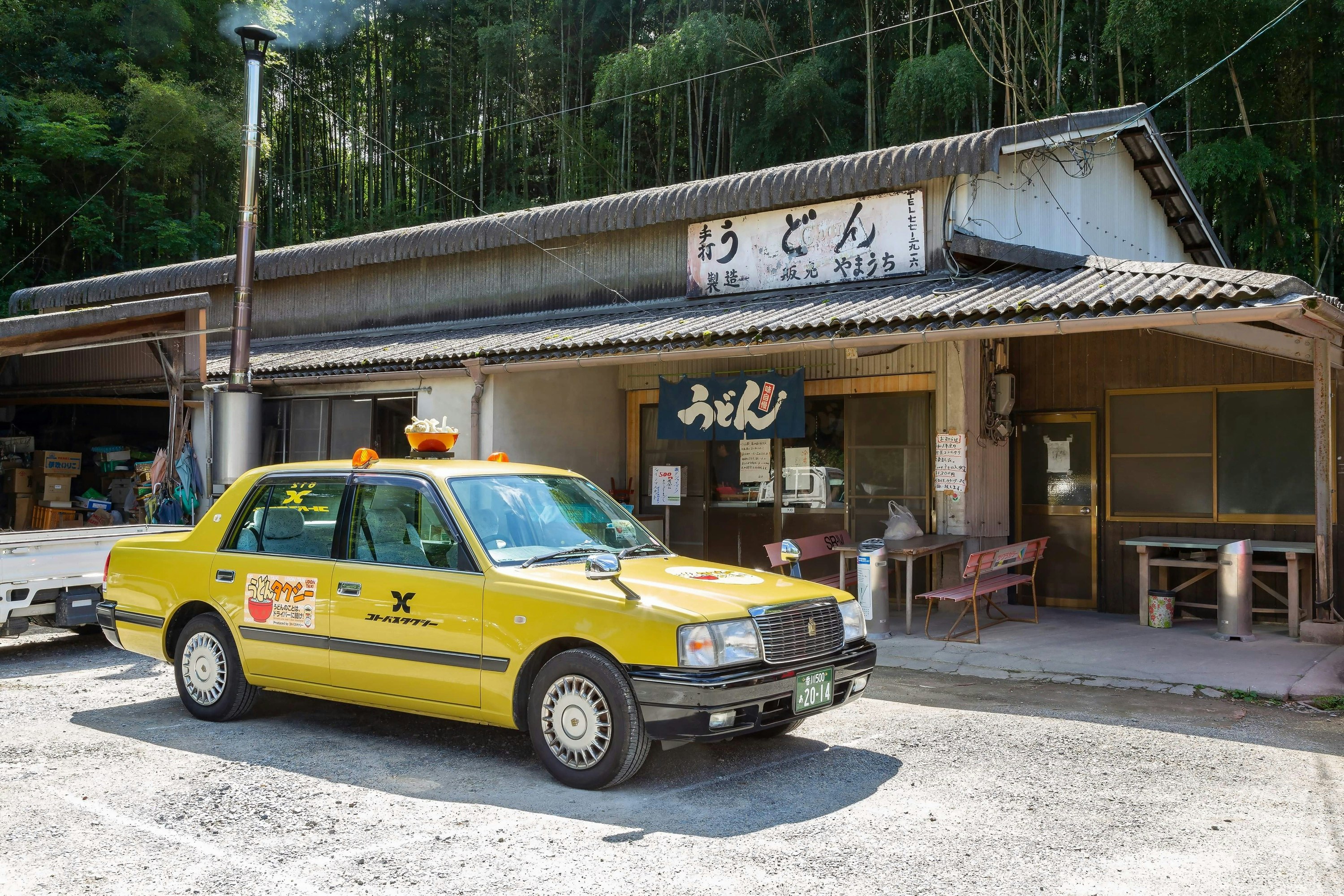 A yellow sedan taxi cab with the name of the taxi company emblazoned on the door in black and a sticker showing a cartoon ramen bowl with the noodles forming letters and a large 3D bowl of ramen on the roof where a police siren would be sits in front of a noodle stand with a tile roof in Kagawa, Japan