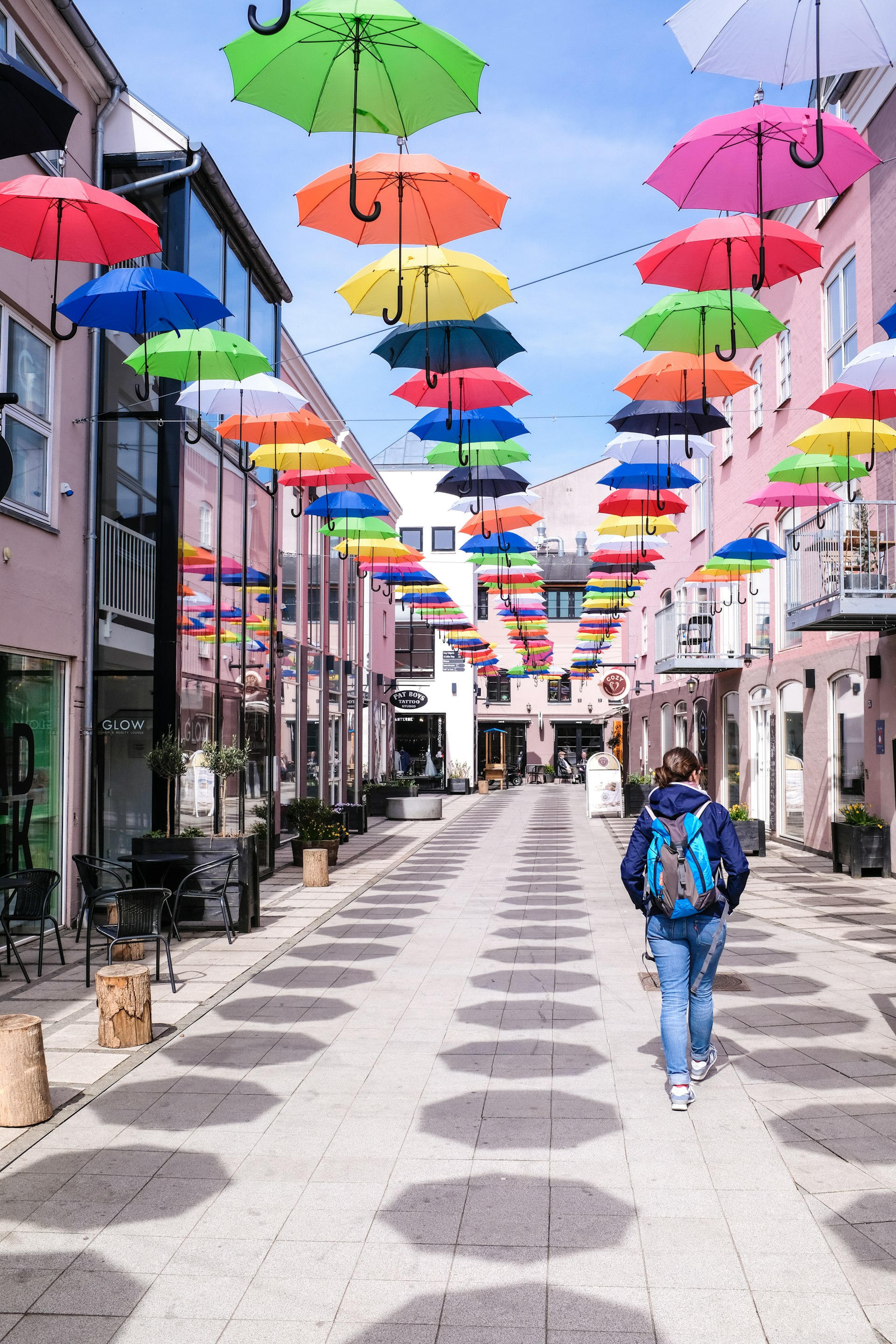 A woman is walking down a pedestrian street in Vejle, Denmark. Brightly coloured umbrellas are hanging overhead and casting octagonal shadows on the street.