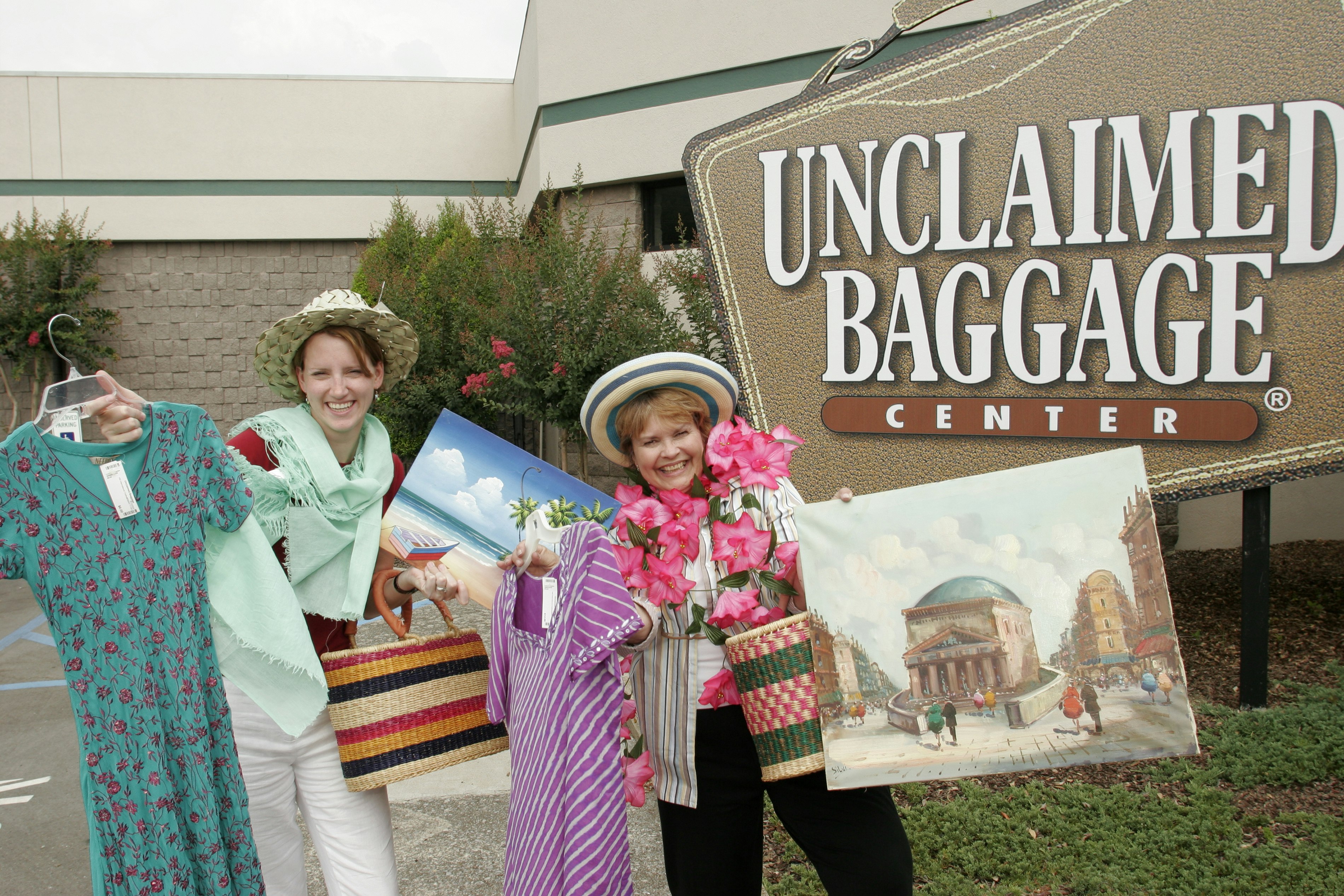 Two women stand by the Unclaimed Baggage Center sign which is brown and suitcase shaped. The woman on the left holds a green print dress on a hangar, a striped wicker bag, and a painting of the beach. On her head is a hat and around her neck is a mint green scarf. The woman on the right also wears a straw hat, and is holding a pink striped dress, a wicker bag, and a painting of a cityscape.