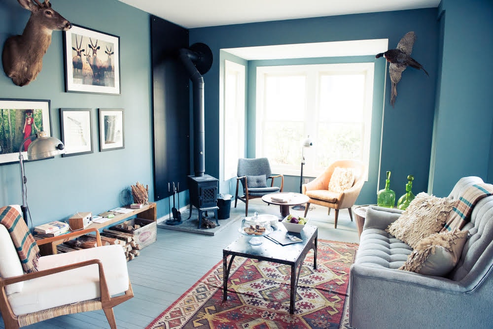 A cozy, soft blue room decorated with neutral-toned midcentury modern style furniture, a small cast iron wood stove, a red, yellow and white tone Kilim rug, and a taxidermic deer head and a pheasant posed as if in flight