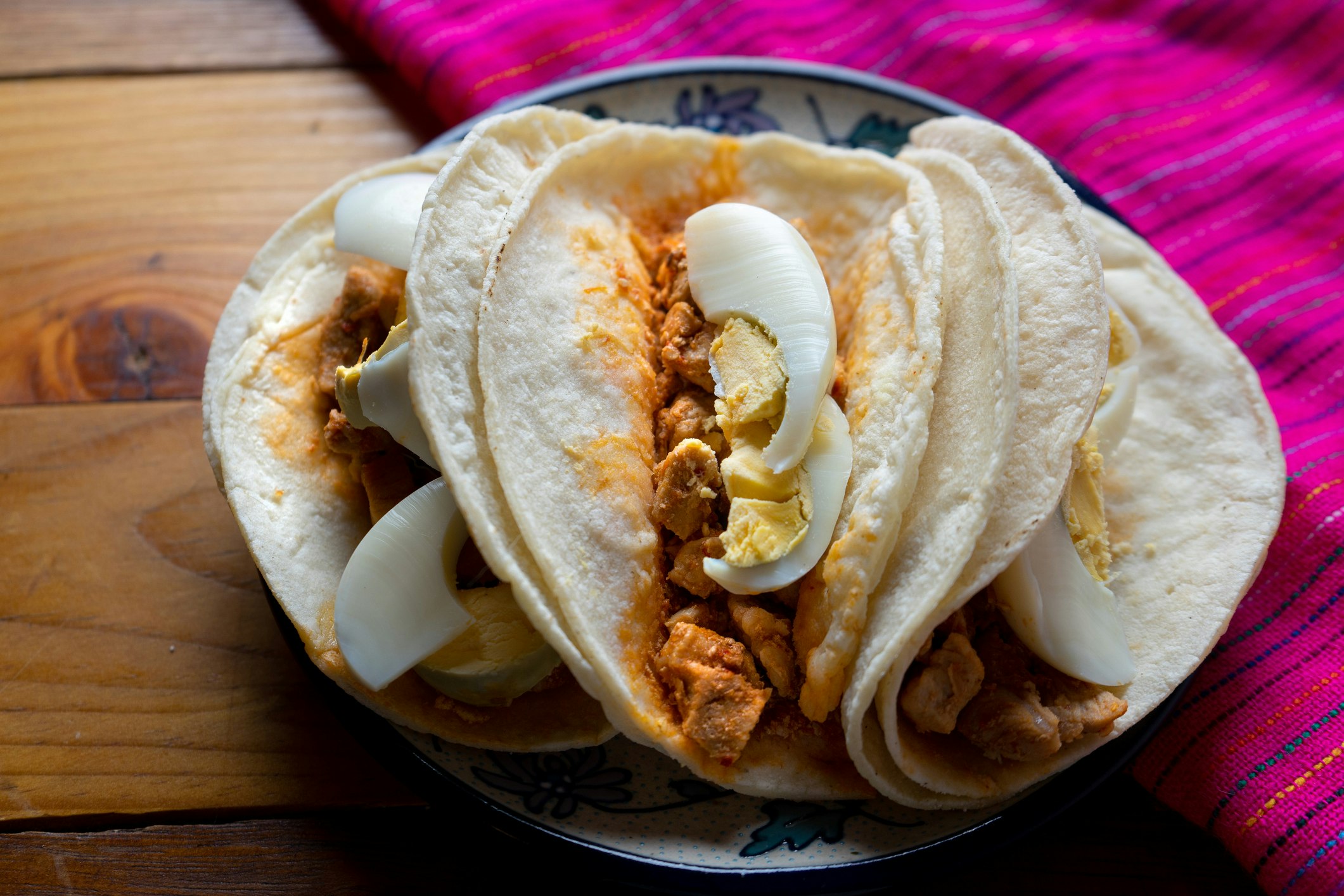 A blue and white plate of three pork tenderloin tacos topped with hardboiled egg in double flour tortillas sits on a warm wooden table next to a hot magenta woven cloth with tiny blue and yellow stripes
