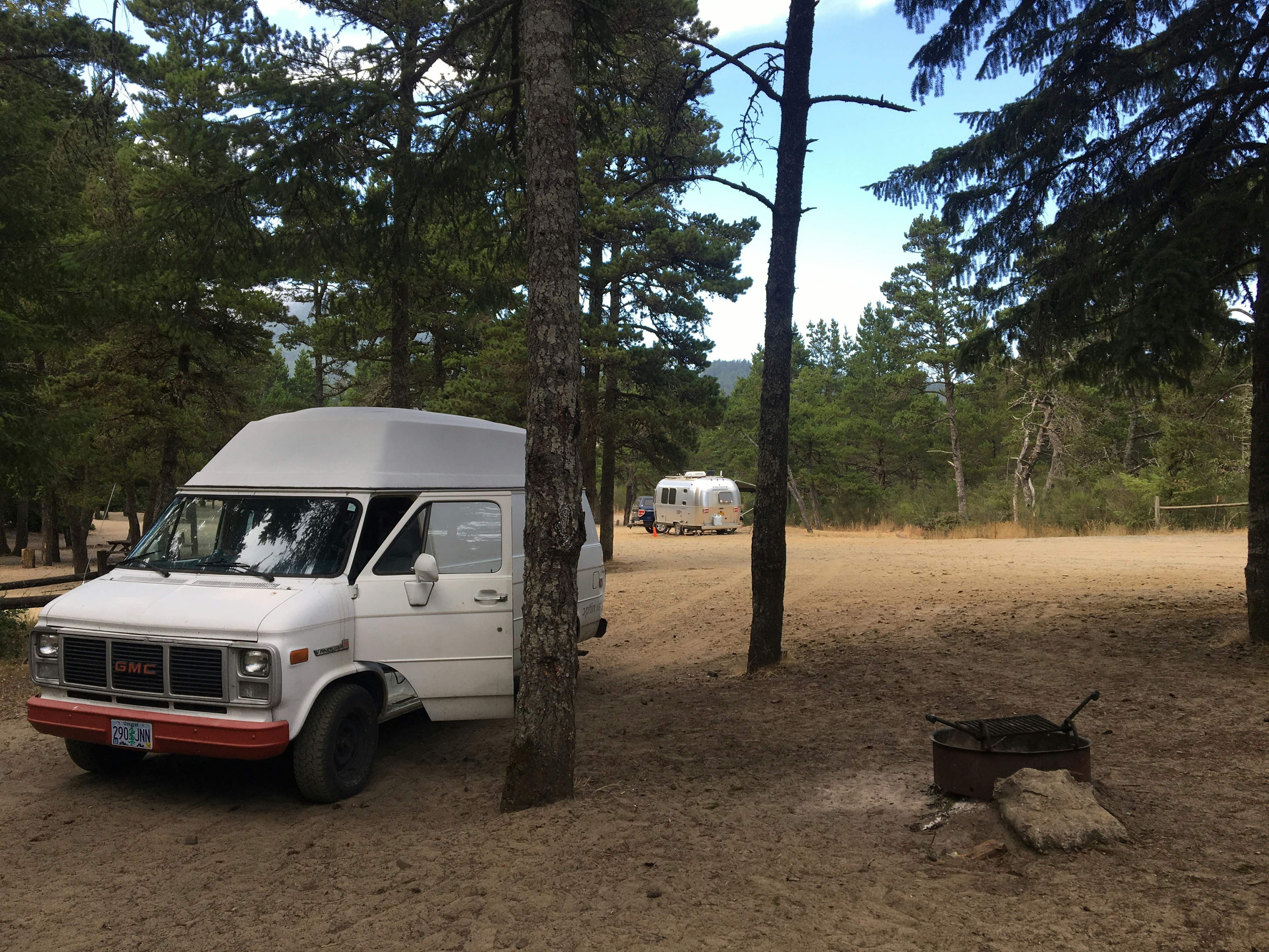 A white van with a red bumper and GMC logo on the grill sits at a campsite amongst pine trees. A metal firepit ring with grill grate sits to the right of the van. In the background is a silver Airstream trailer at another campsite.