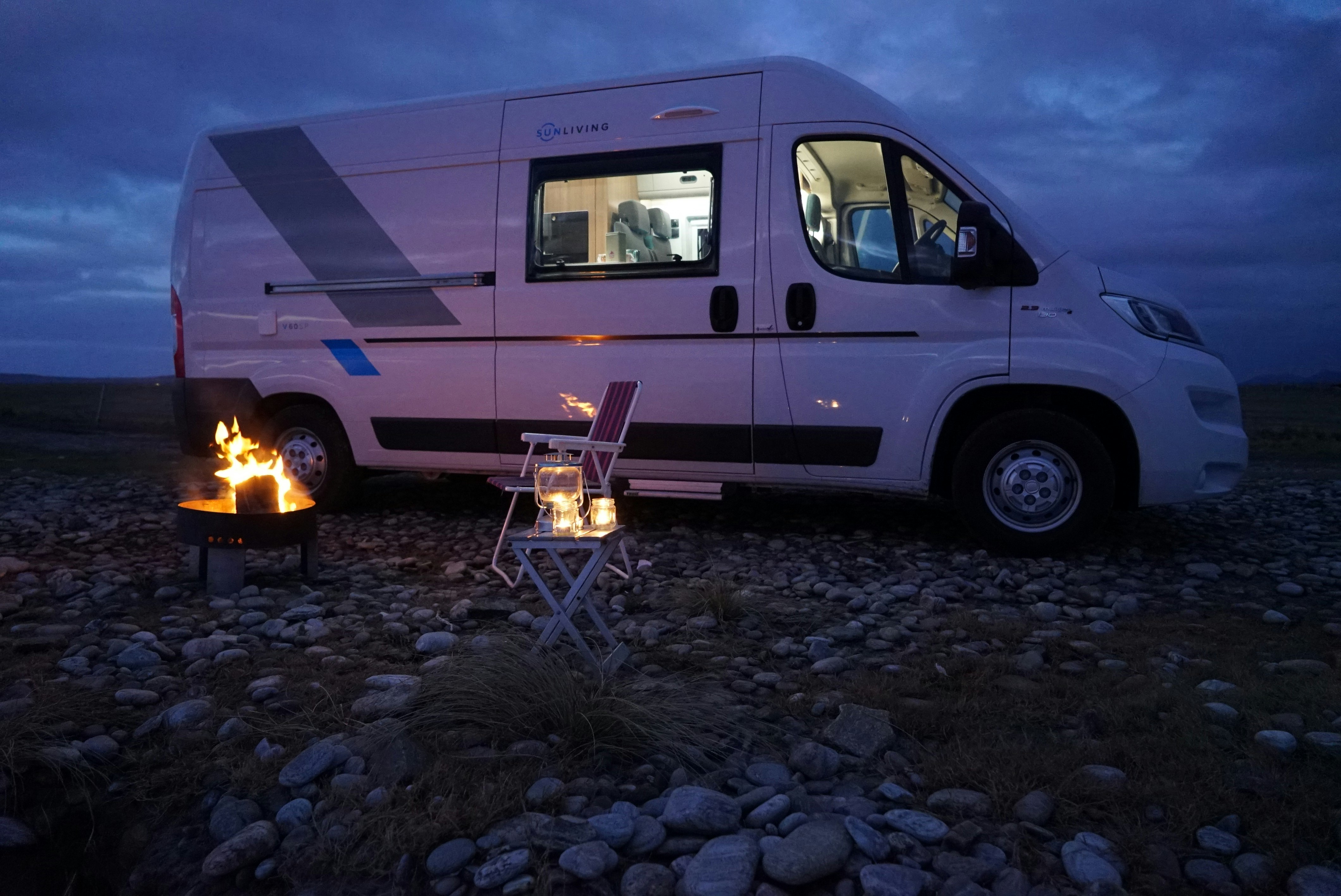 A large white van is parked in the middle of the wilderness. Next to it, a lone camping chair stands next to a small camp fire.