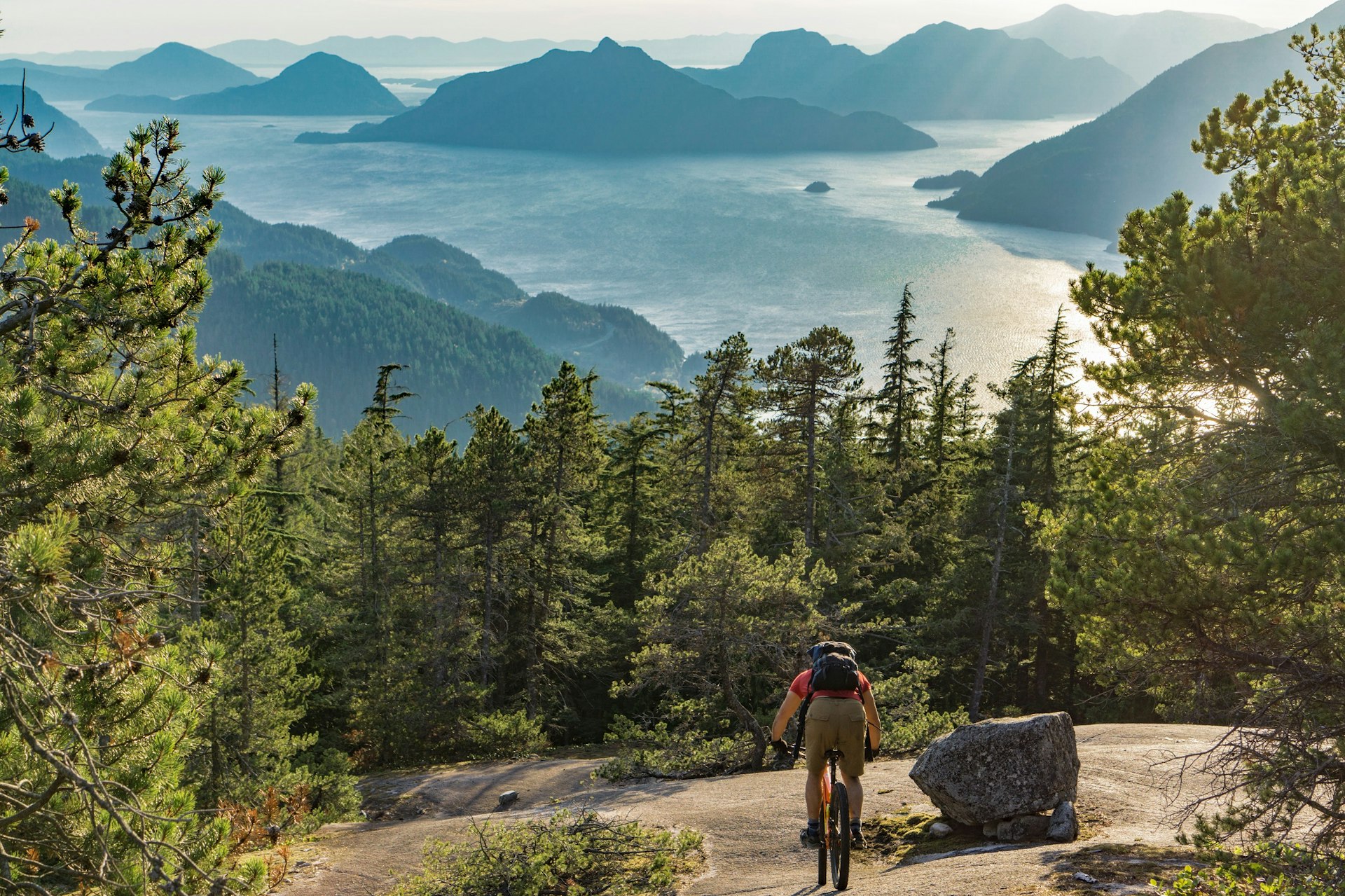 A mountain biker descends down a massive granite outcrop towards the forest below; in the distance far below is a spellbinding scene of forested islands jutting out of the ocean.