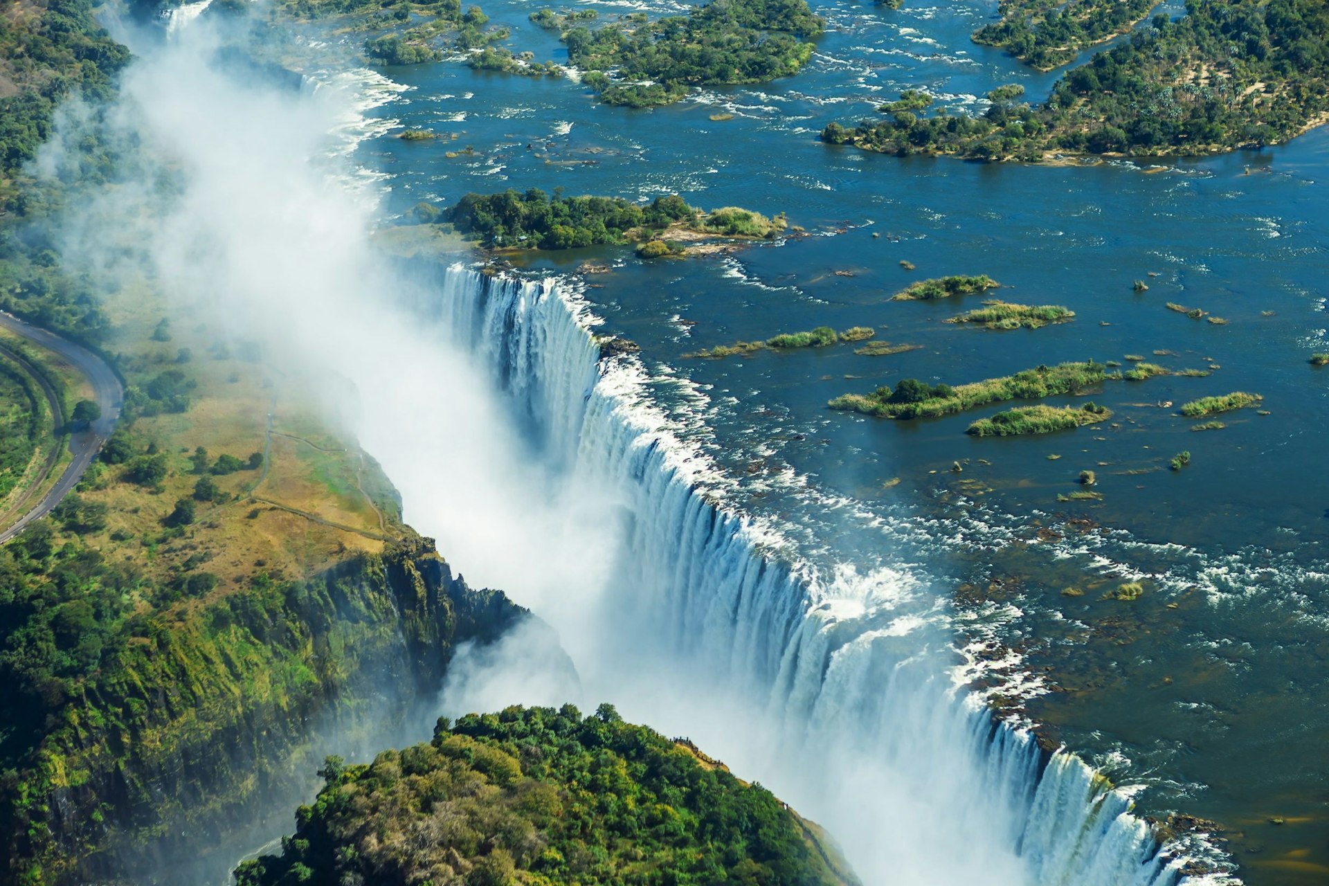 A curtain of water plunges over the mile-wide precipice that is Victoria Falls; misty clouds rise into the skies, while the waters of the Zambezi are channeled through the narrow gorge that makes the border between Zimbabwe and Zambia.