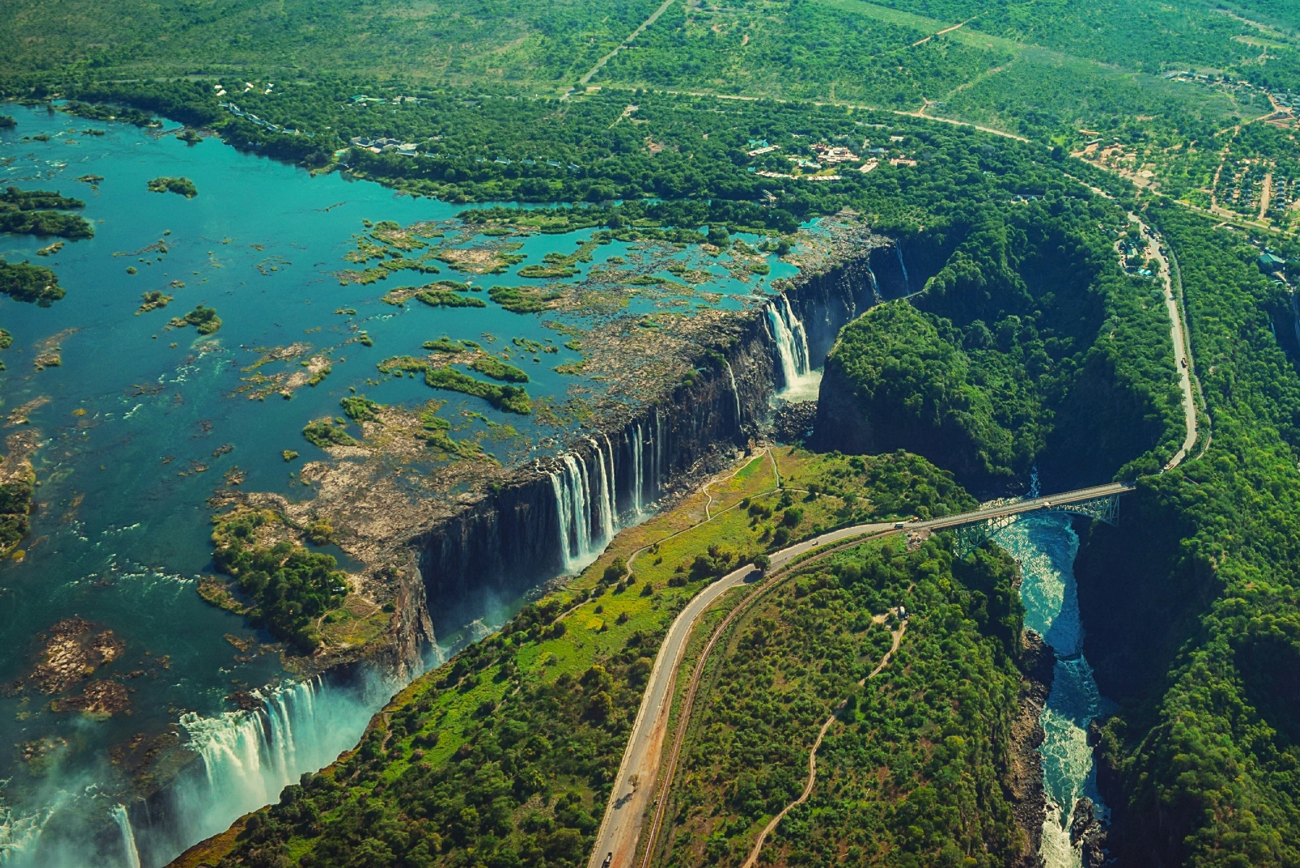 An aerial shot shows lush green and turquoise vegetation around the wide mouth of the Zambezi River where it spills over Victoria falls and winds through a basalt gorge