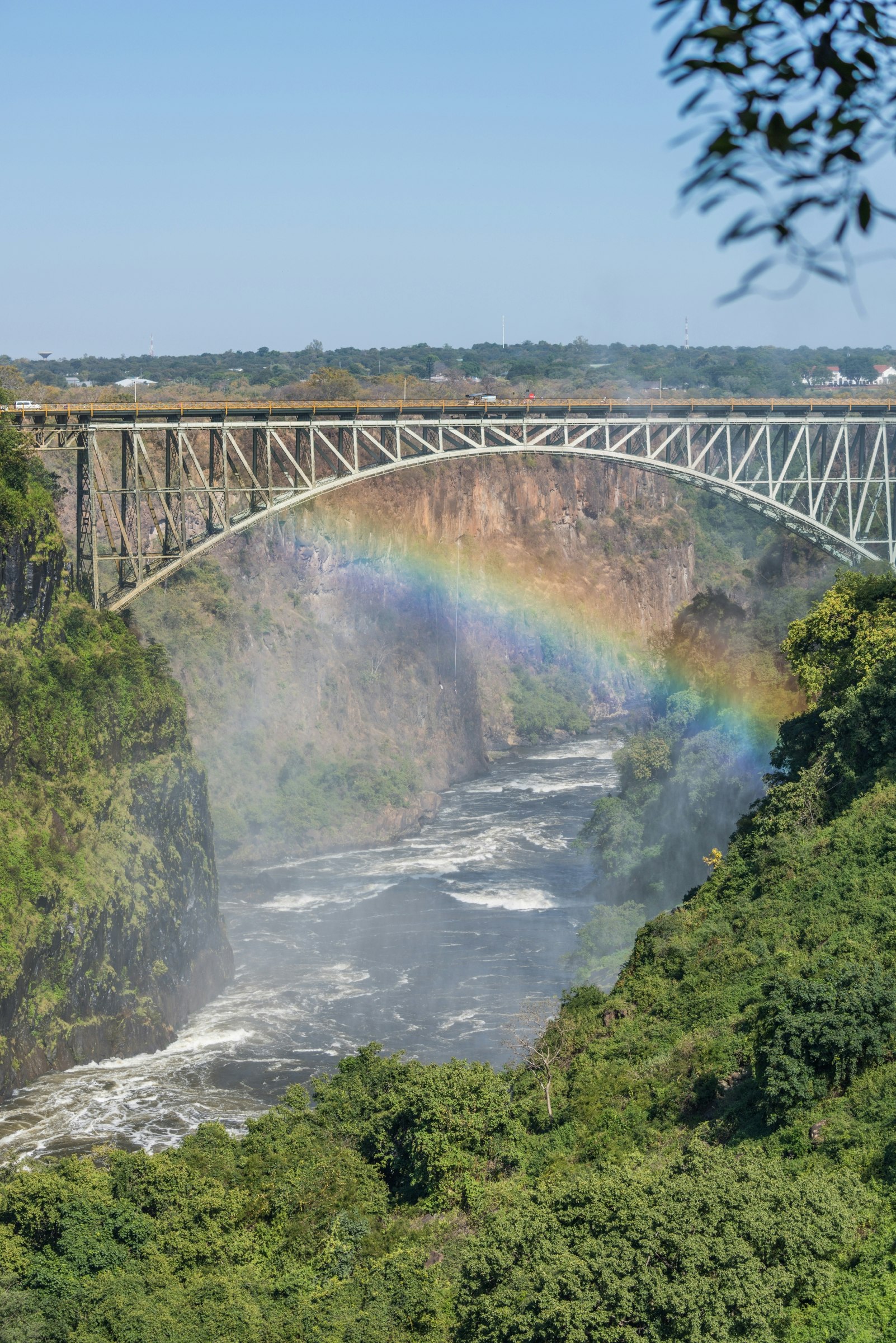 Beyond a foreground of lush bush are the rapids of the Zambezi river and mist from Victoria Falls; behind the rainbow is the graceful arch of Victoria Falls Bridge.