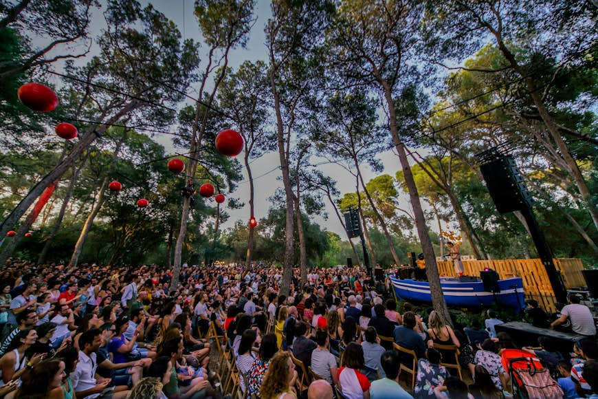 A large crowd of people sitting amidst tall trees, watching a performer at Vida Festival in Spain.