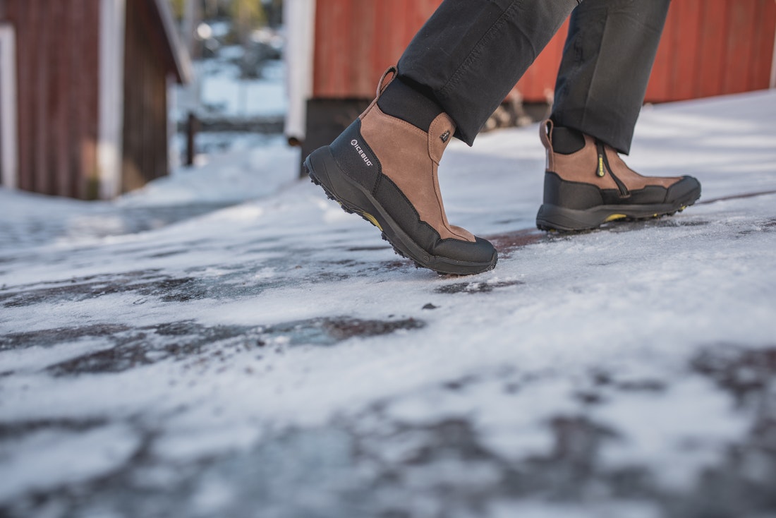 A person in charcoal colored jeans and brown and black Icebug boots walks up a slope slick with snow and ice in front of red clapboard buildings.