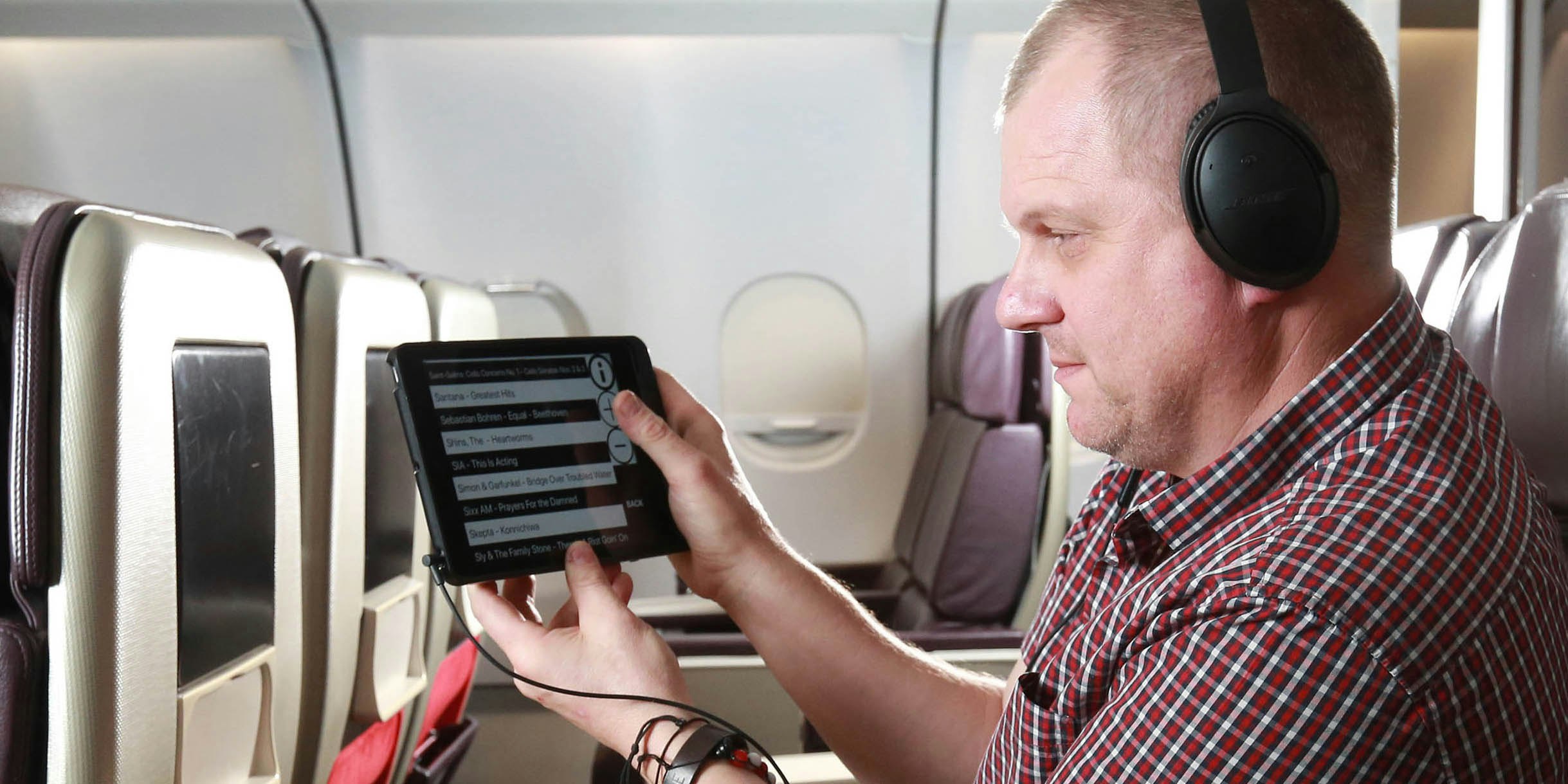 A man sitting in an airplane has his headphones plugged into an iPad. On the screen, the text is large and in black and white contrast.
