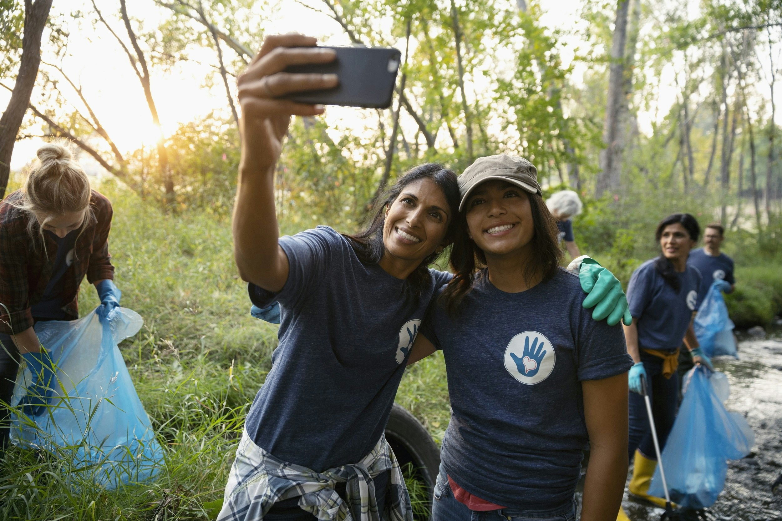 A woman and a teenage girl wearing matching t-shirts pose for a selfie while working with a group clearing litter in a woodland setting