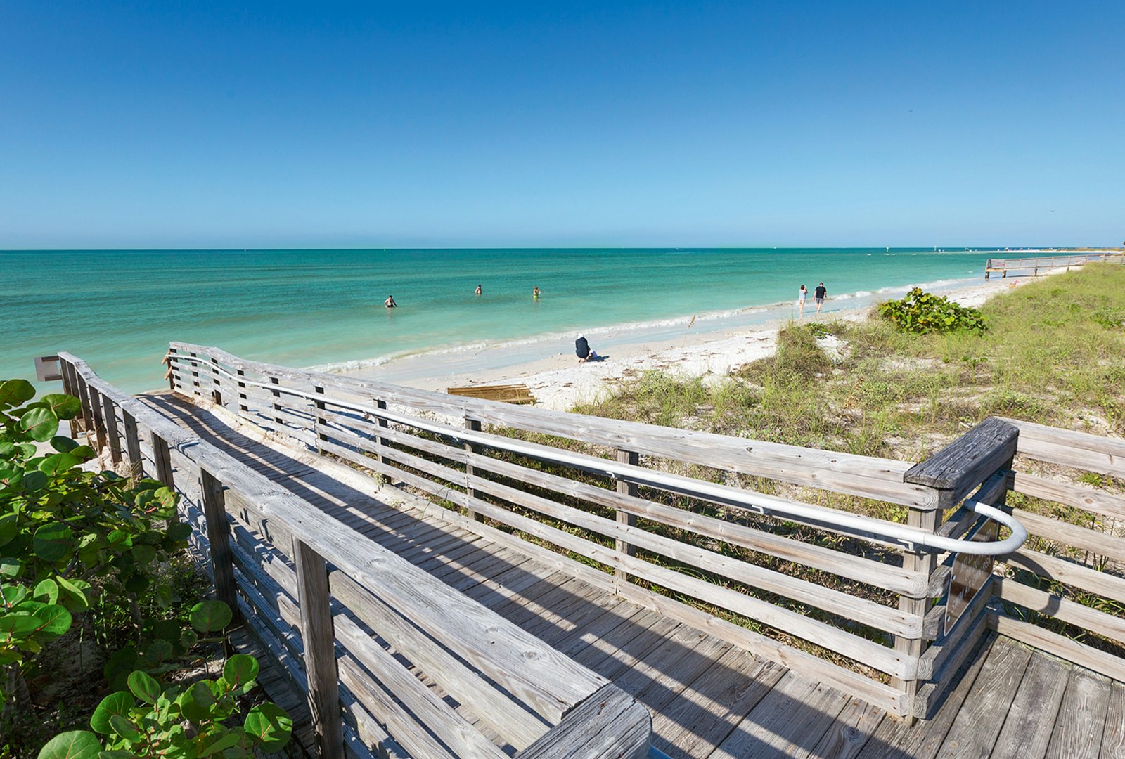 A wood path leads to a white sand beach where beachgoers stand waist deep in turquoise water on Honeymoon Island