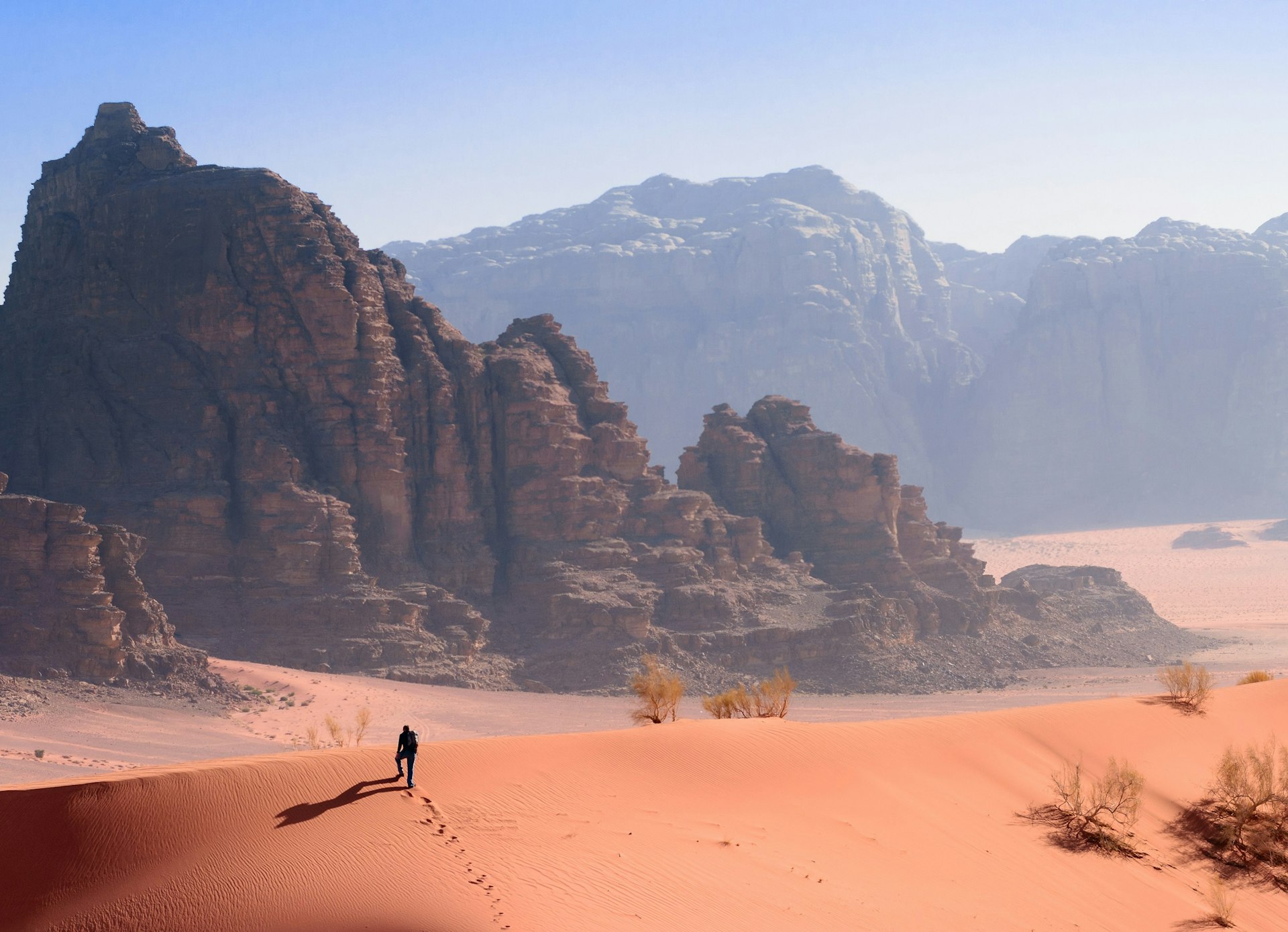 A person hikes up a red sand dune, with the most stunning backdrop in the distance; two sets of towering rocky mountains stand one in front of the other.