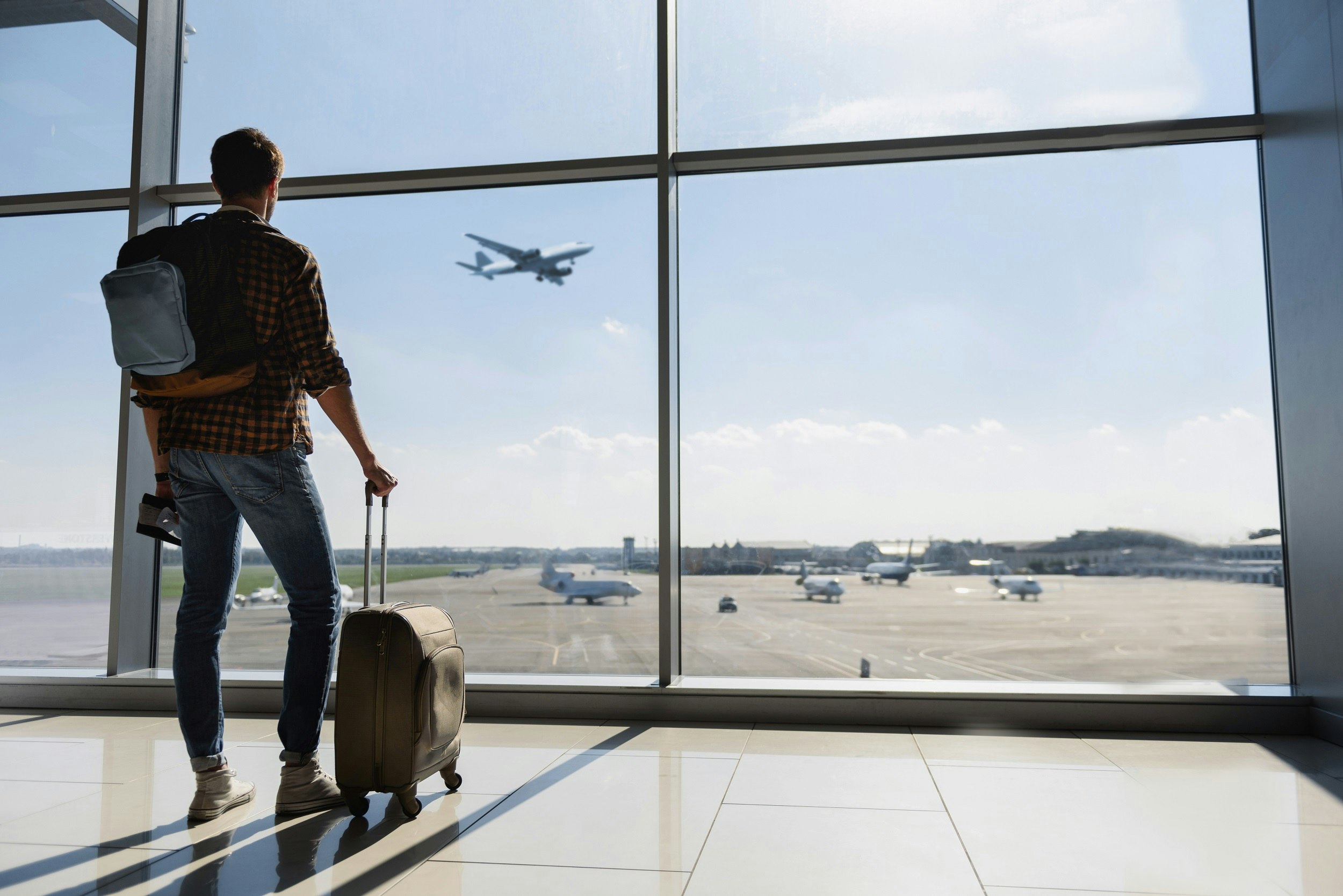 Man with backpack and small case stands looking out the airport window at planes taxiing as a jet flies overhead