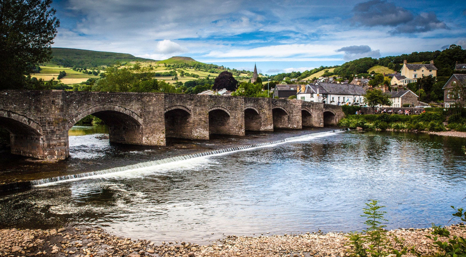 A long stone bridge stretches across a river. In the background are rolling green hills, blue sky and the top of a church steeple.