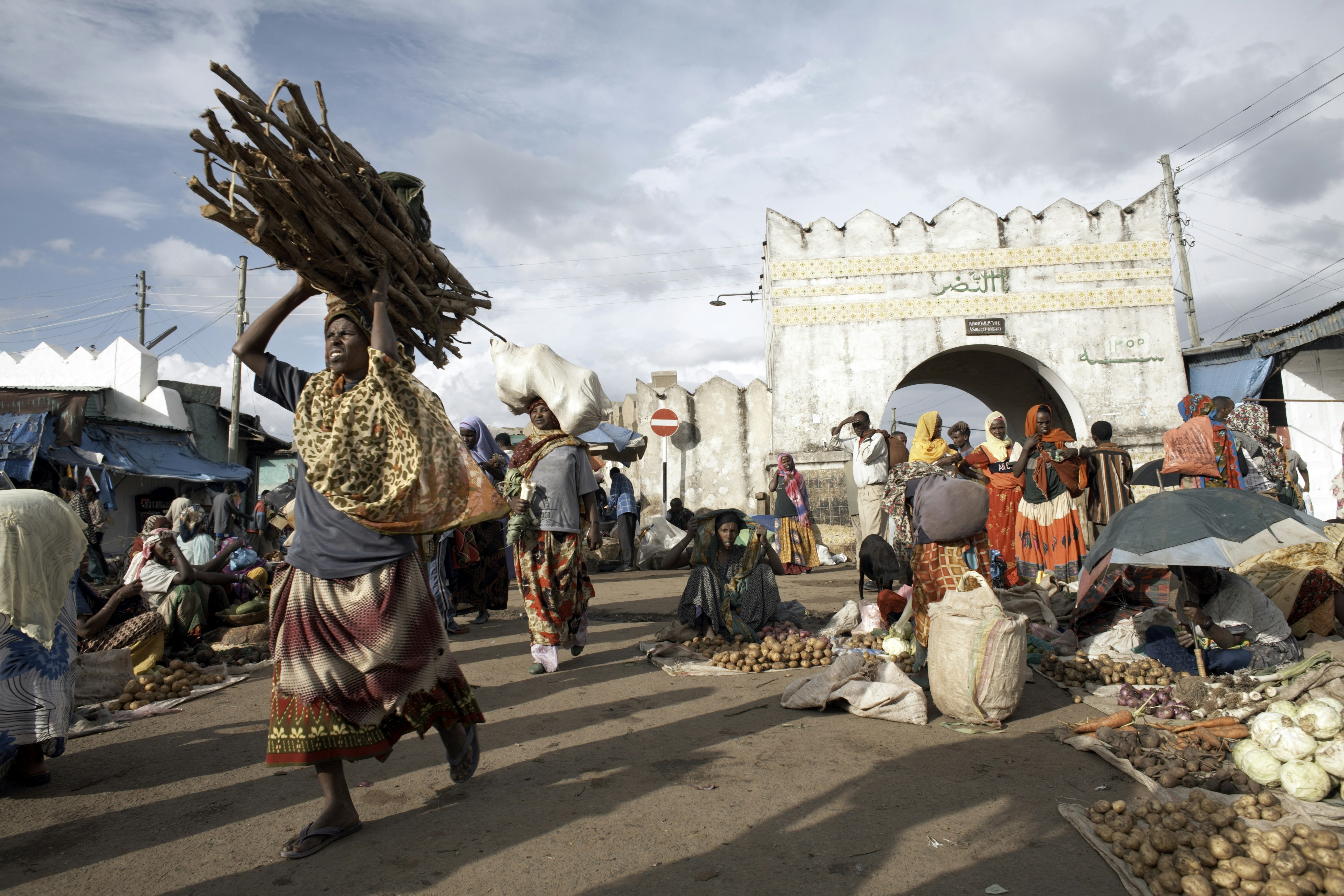 A busy market scene close to the large white stone gate leading into Harar Old Town. Many locals are carrying goods and produce.