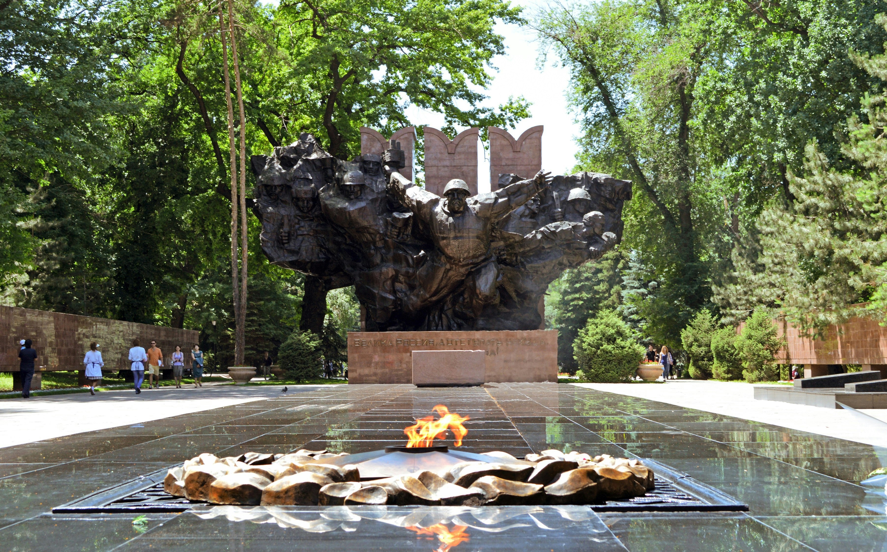 A large black war monument - showing soldiers bursting from a map of the USSR - stands behind a small fire pit; an eternal flame honouring solders who lost their lives in conflict. Behind the monument, trees and other greenery is visible.