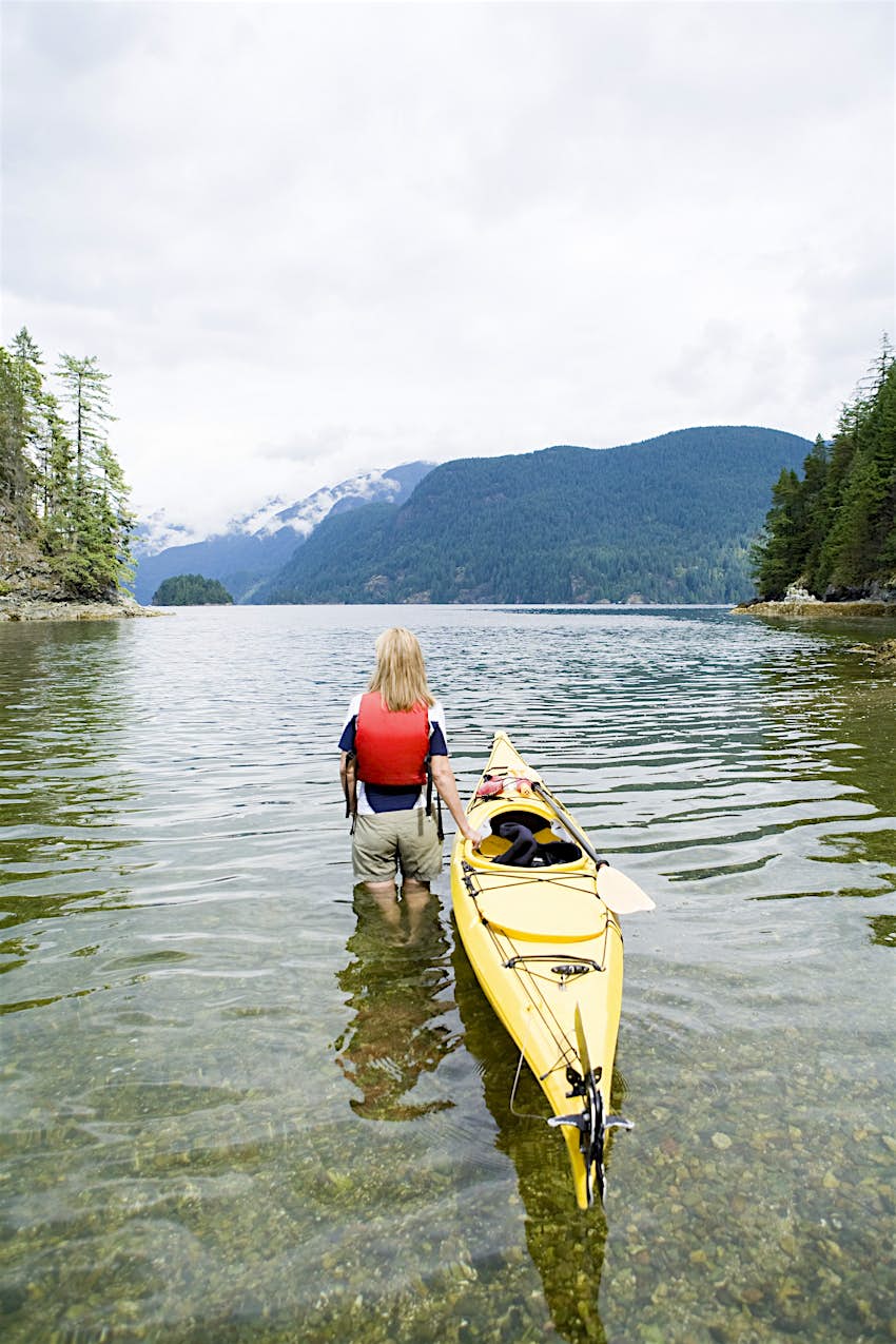 watery-escape-indian-arm-vancouver A woman stands with her back to the camera in water that is up to her knees; she holds a kayak and looks out to the forested fjord of Indian Arm.