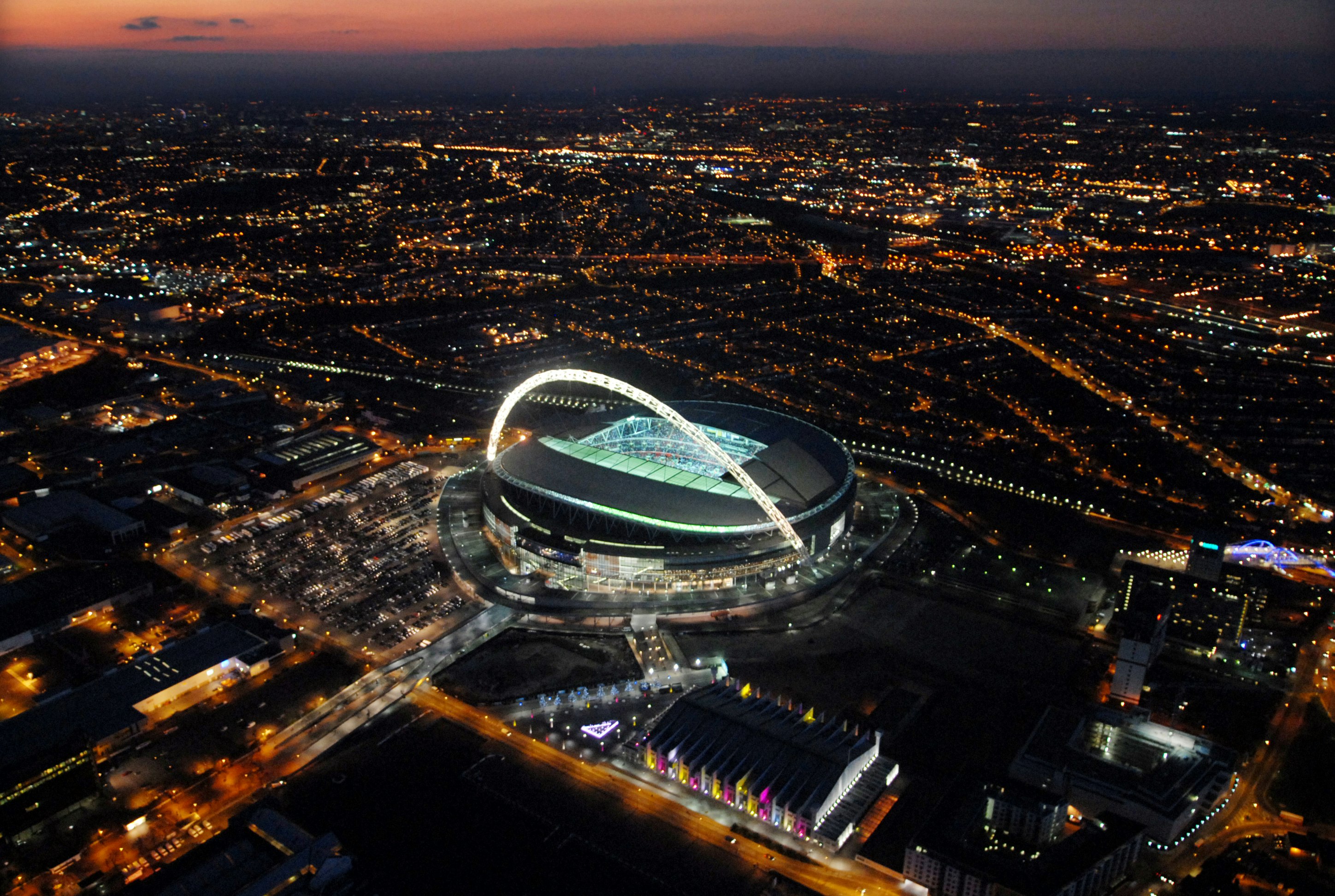 An aerial shot of London's Wembley Stadium at night; the stadium's famous arch is illuminated bright white.