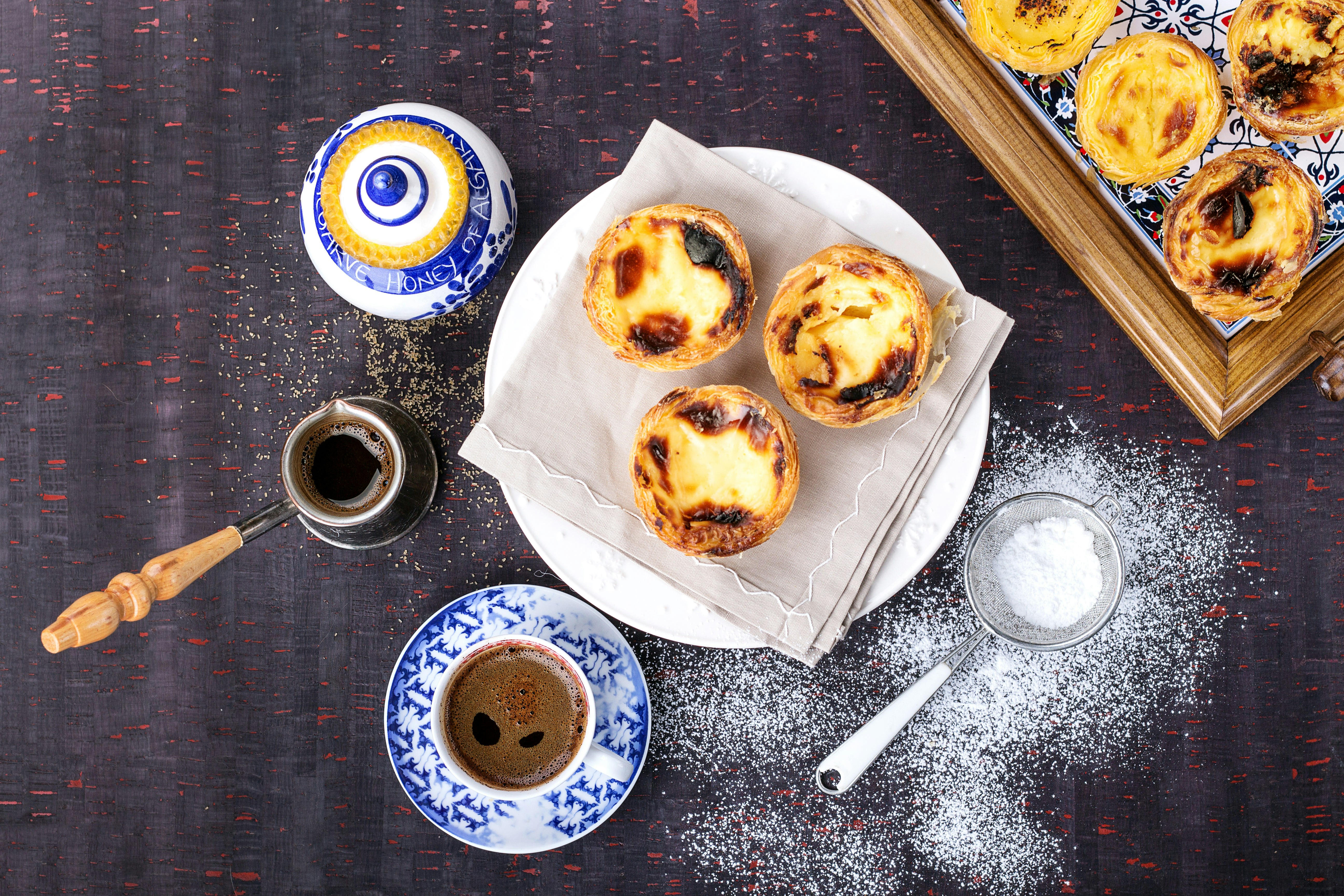 Traditionell portugisisk bakelse Pastel de Nata serveras med kaffe och honung på en bricka över en rustik träskiva.  Det finns även en sil med florsocker bredvid bakverken på brädan.