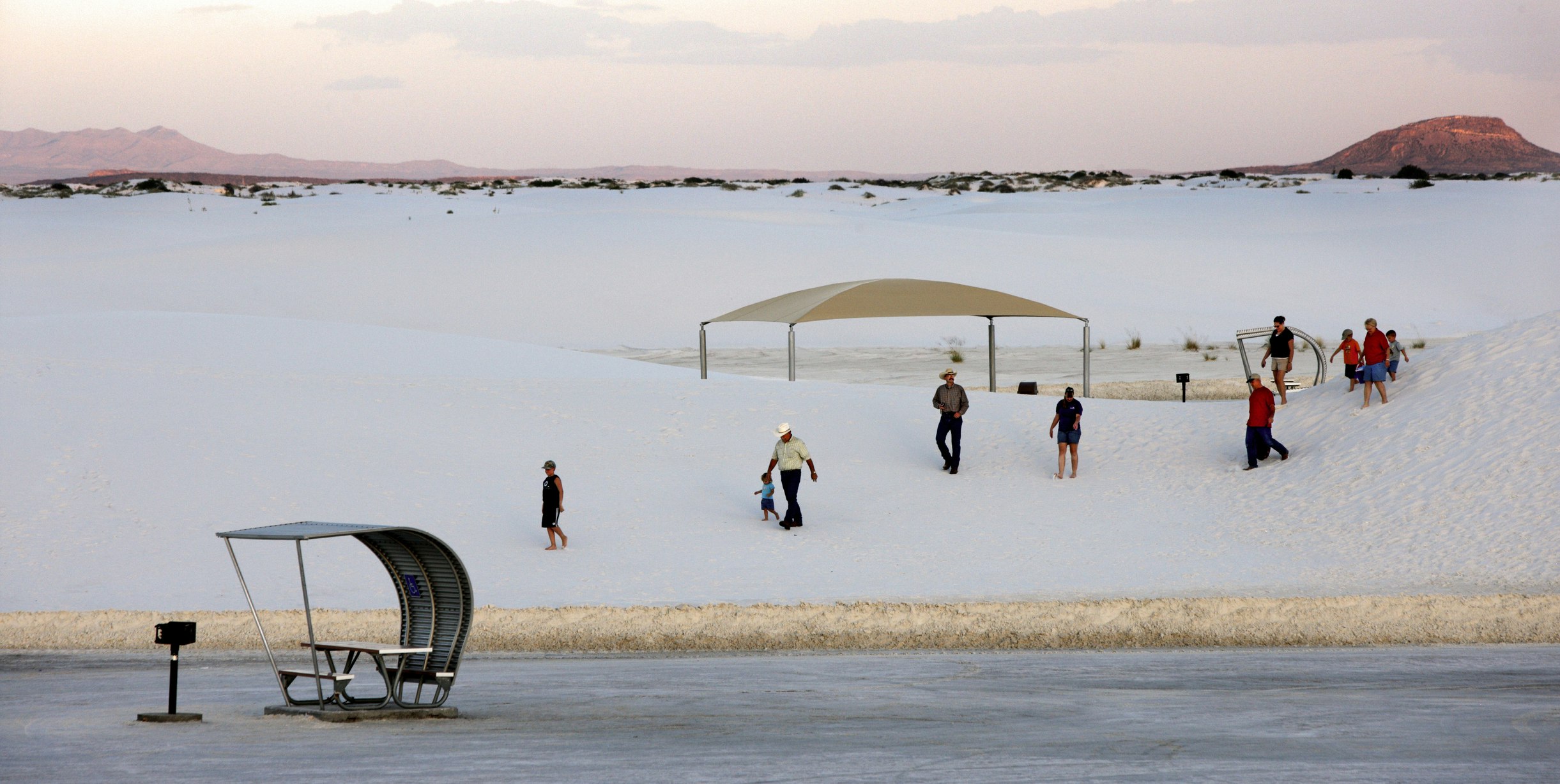 A stream of visitors cross into the parking lot at White Sands National Monument with a outhouse style bathroom to the left and a silver hooded picnic table dotting the landscape