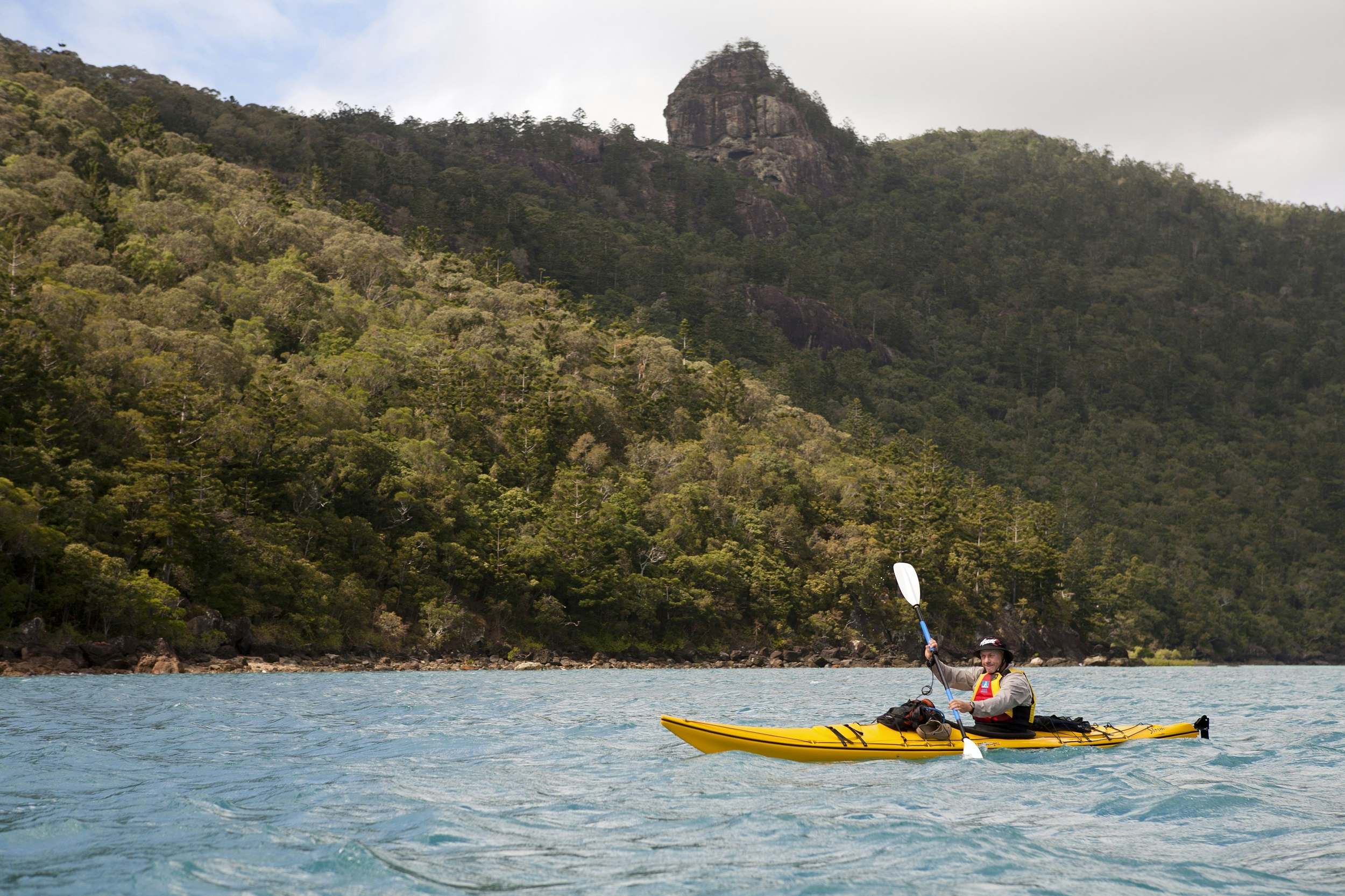 A man paddles a yellow sea kayak across choppy waters with a forested island of the Whitsundays in the background.