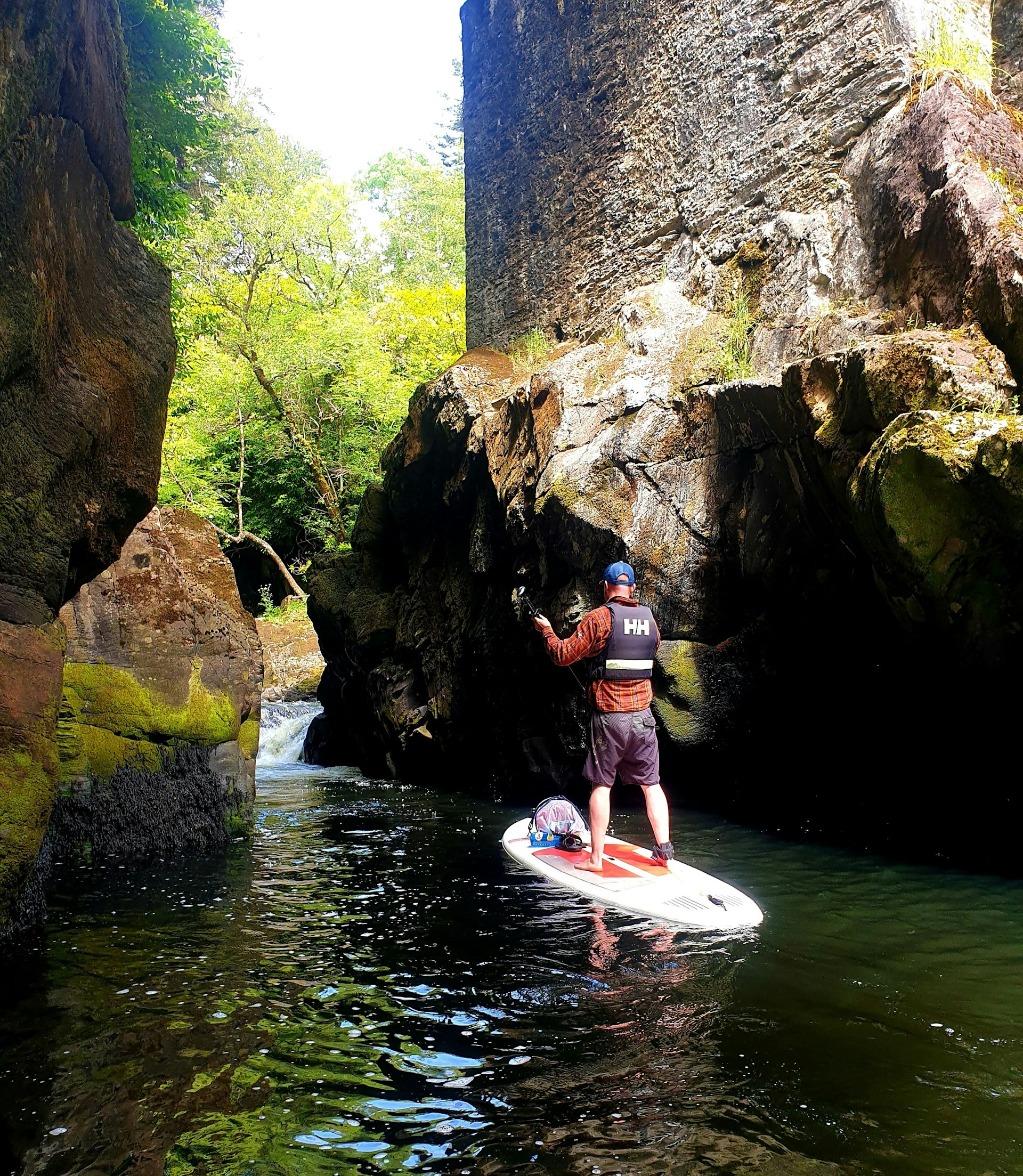 A paddler makes his way on a SUP between a small chasm between two rock walls; a forest looms on the other side.