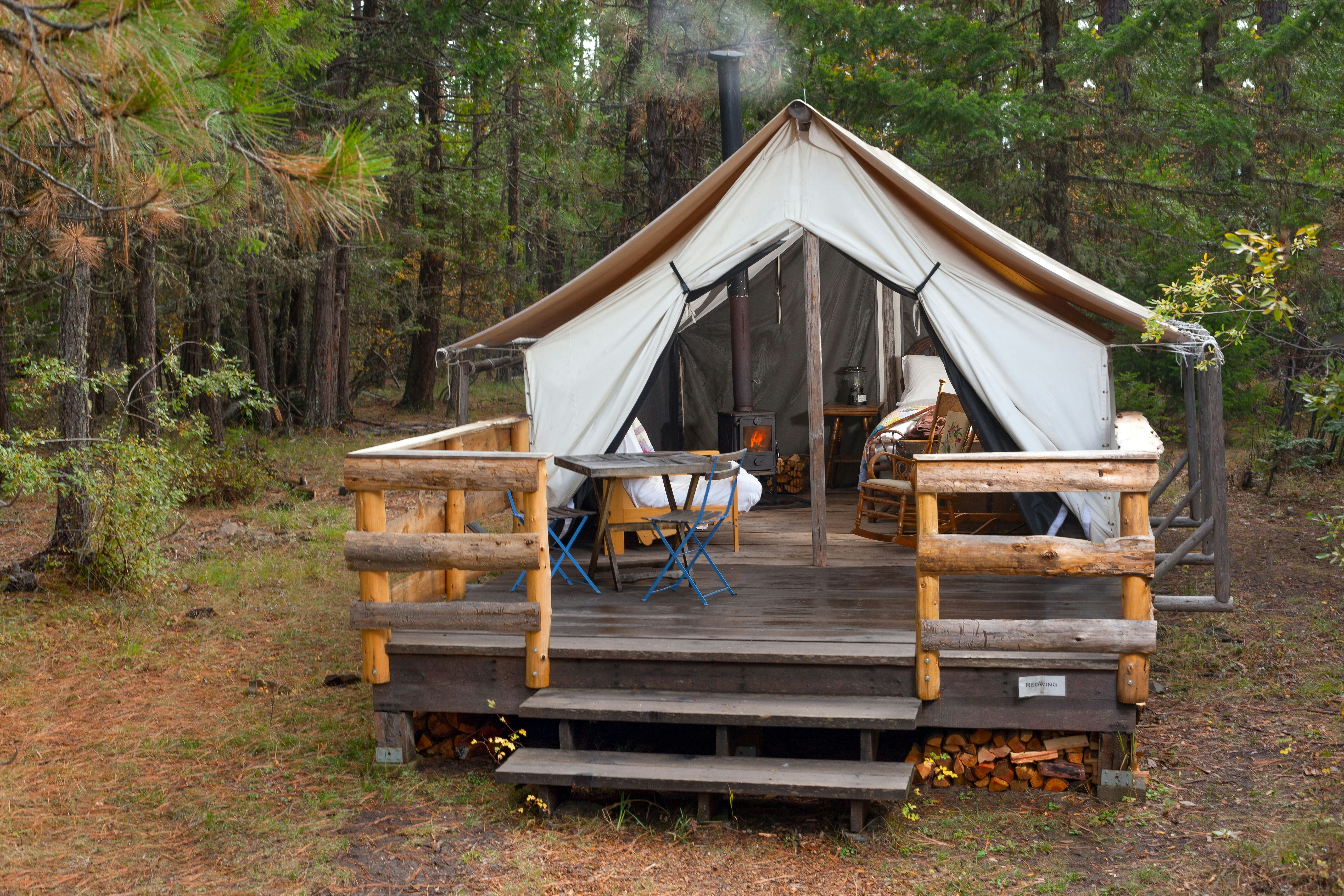 A canvas glamping tent at Willow-Witt Ranch in Ashland, Oregon is outfitted with a woodburning cast iron stove, a vintage rocking chair, and a bed covered with a antique-style quilt. A table with blue folding chairs sits on the porch, formed by a wooden platform on which the tent sits. Logs are stored underneath and a pine forest is all around.