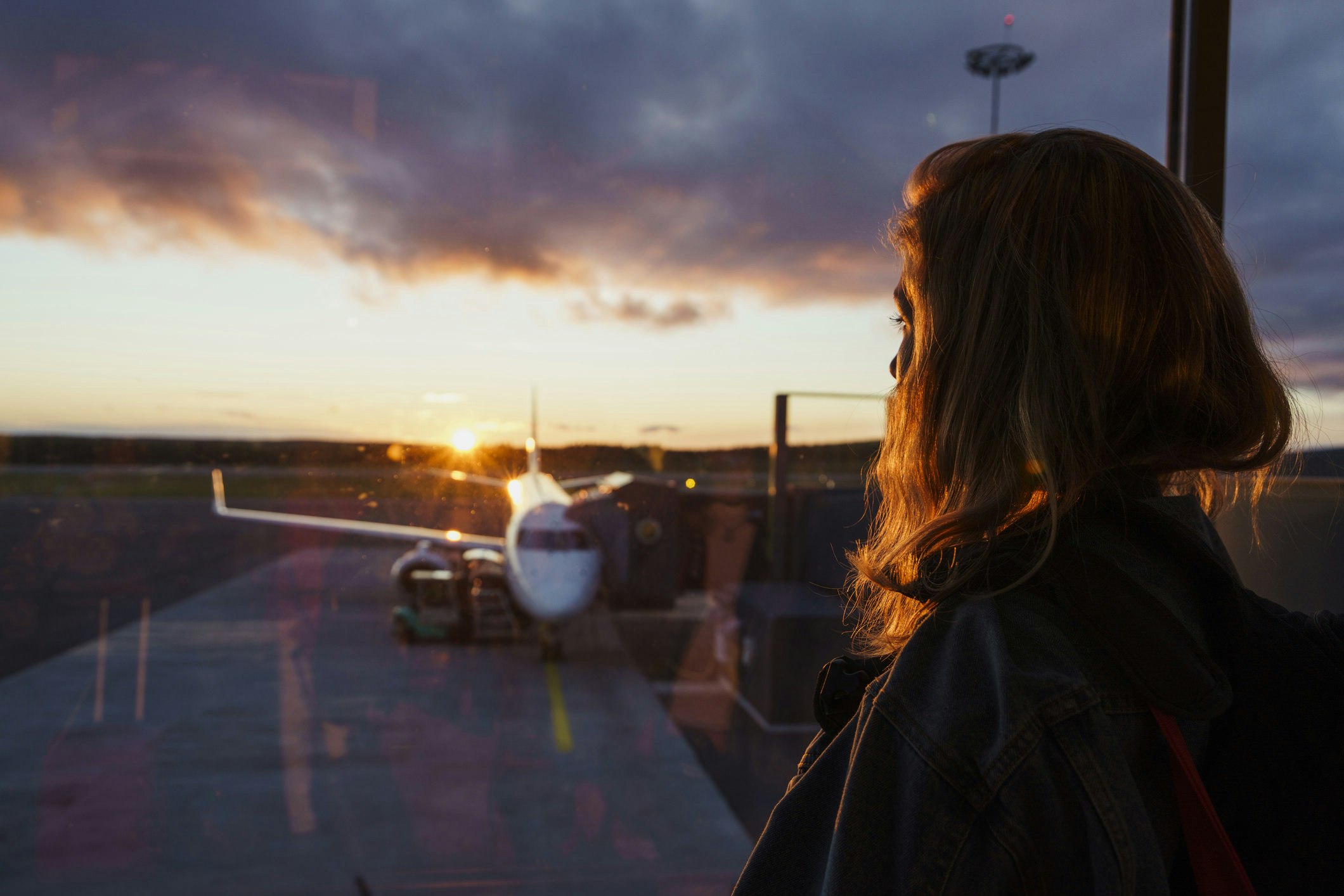 A woman is in the departure lounge, anxiously looking out at a plane as the sun sets behind it.