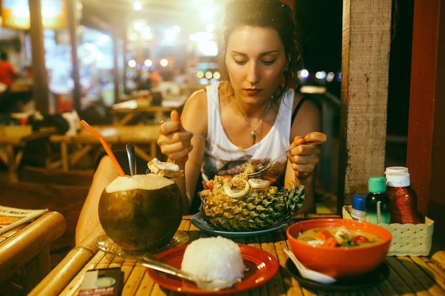 A woman in a white vest eats noodles from a pineapple in a rustic-looking restaurant