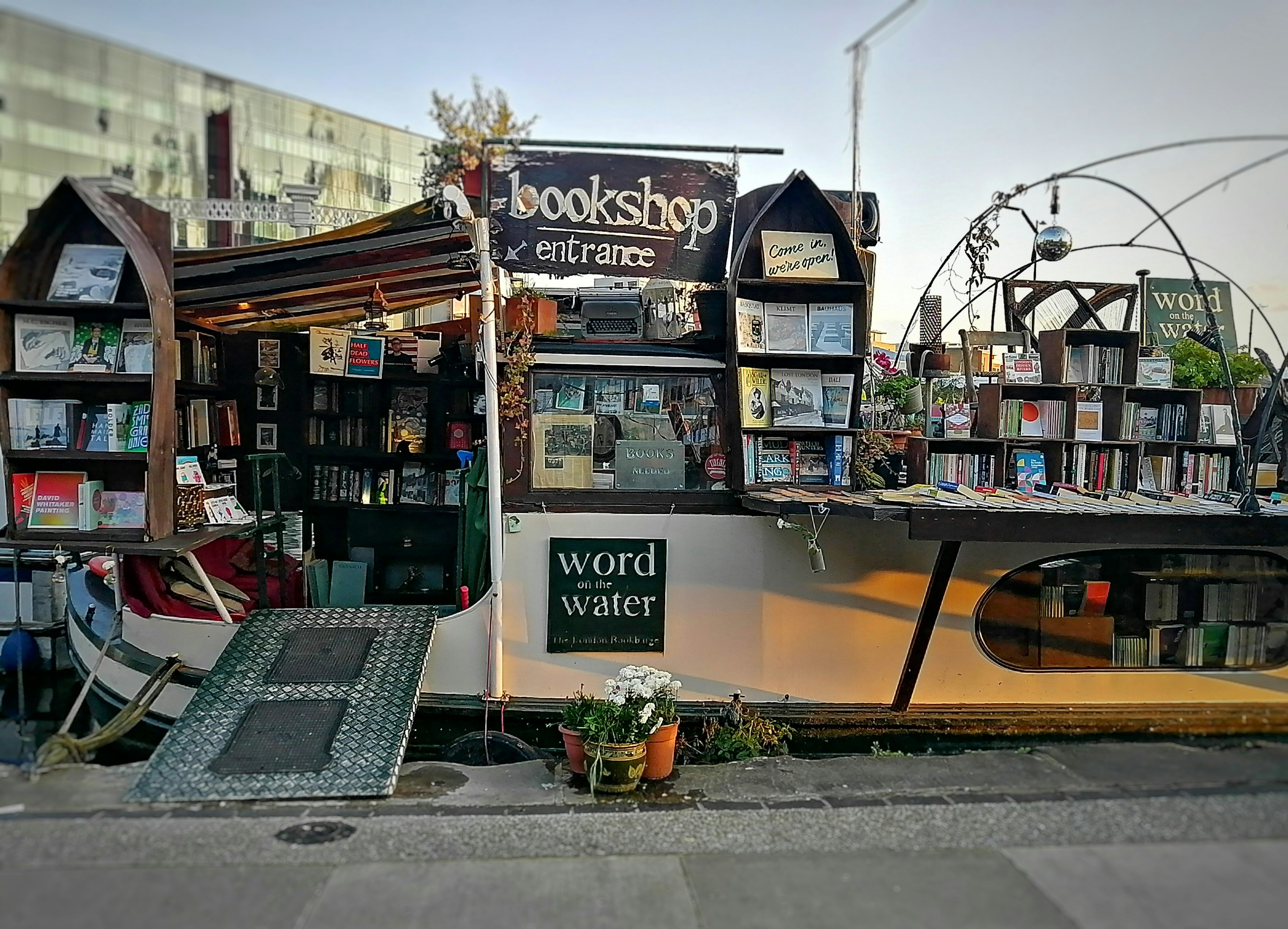 A barge docked by the side of the river. The sides of the barge are covered in shelves that are packed with books. A sign says 'Bookshop entrance' with an arrow pointing to the door