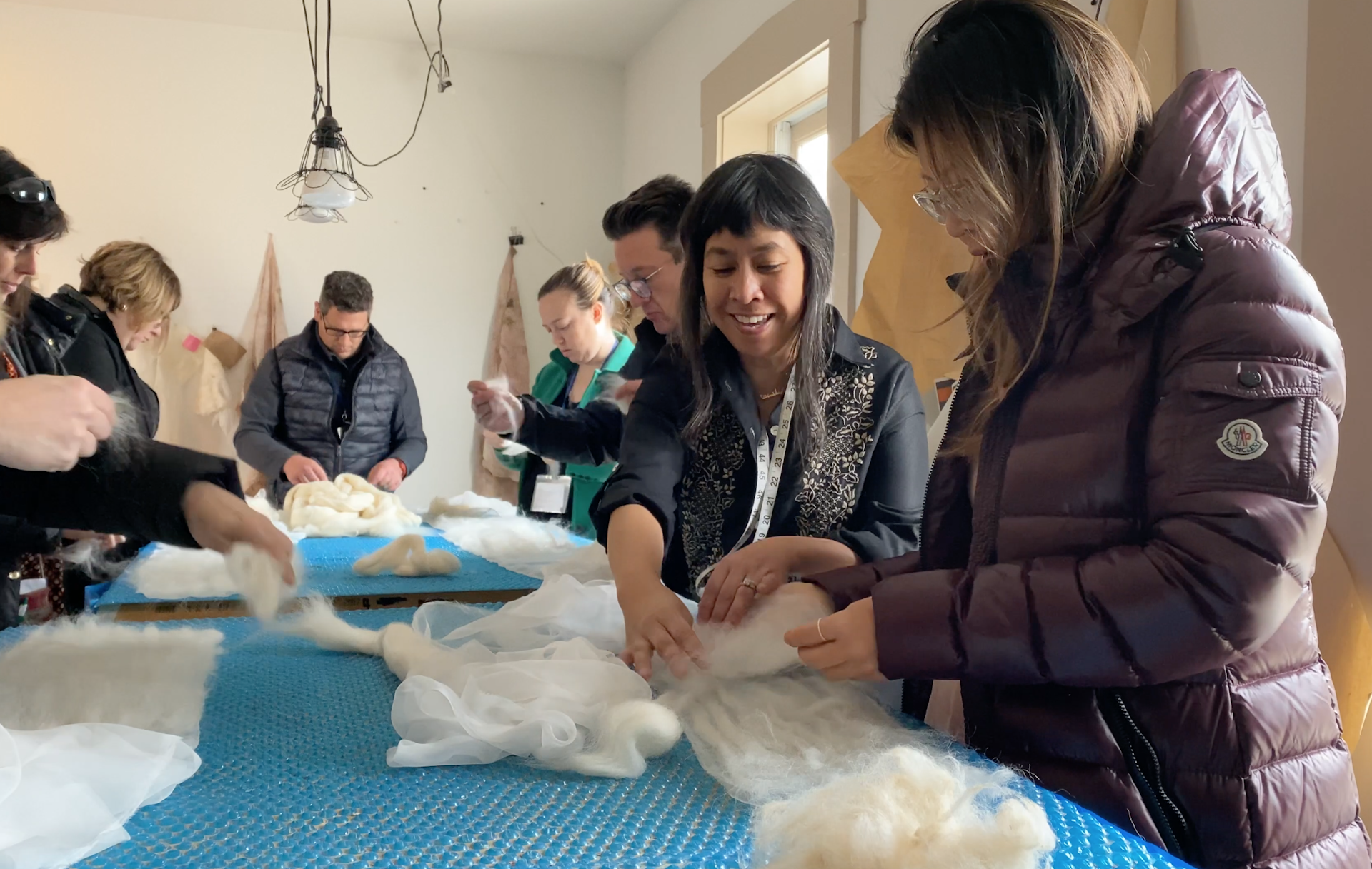 Celeste Malvar-Stewart shows one of her workshop students how to manipulate the wool to create a scarf at her Columbus, Ohio studio called Hangar 391. Seven people stand around a table covered in blue bubble wrap and white wool in an airy, light-filled room.