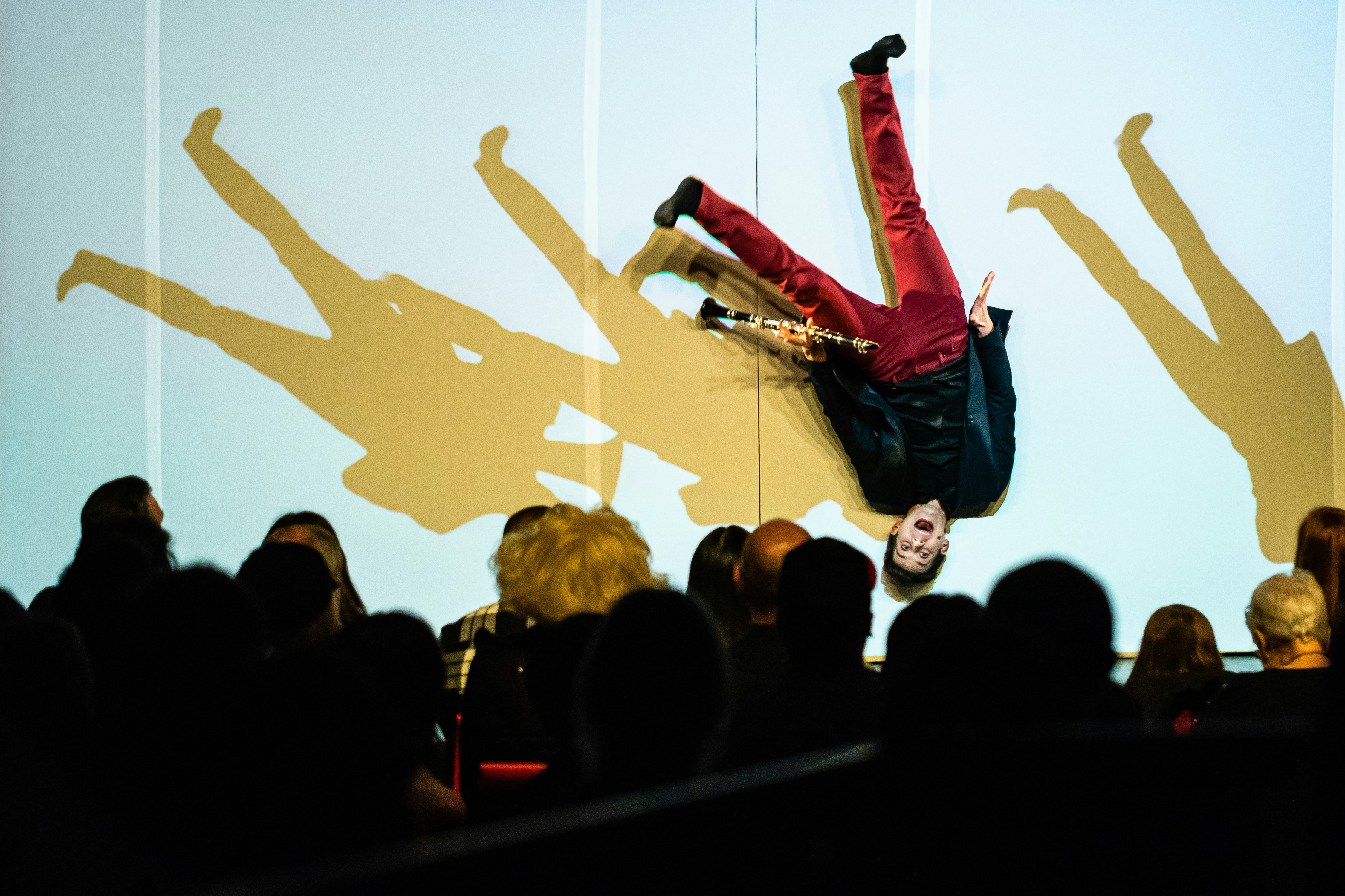 A photo taken from the stalls, as magician Xavier Mortimer flails upside down on stage, holding a clarinet. Four shadows beside him are in a similar pose.