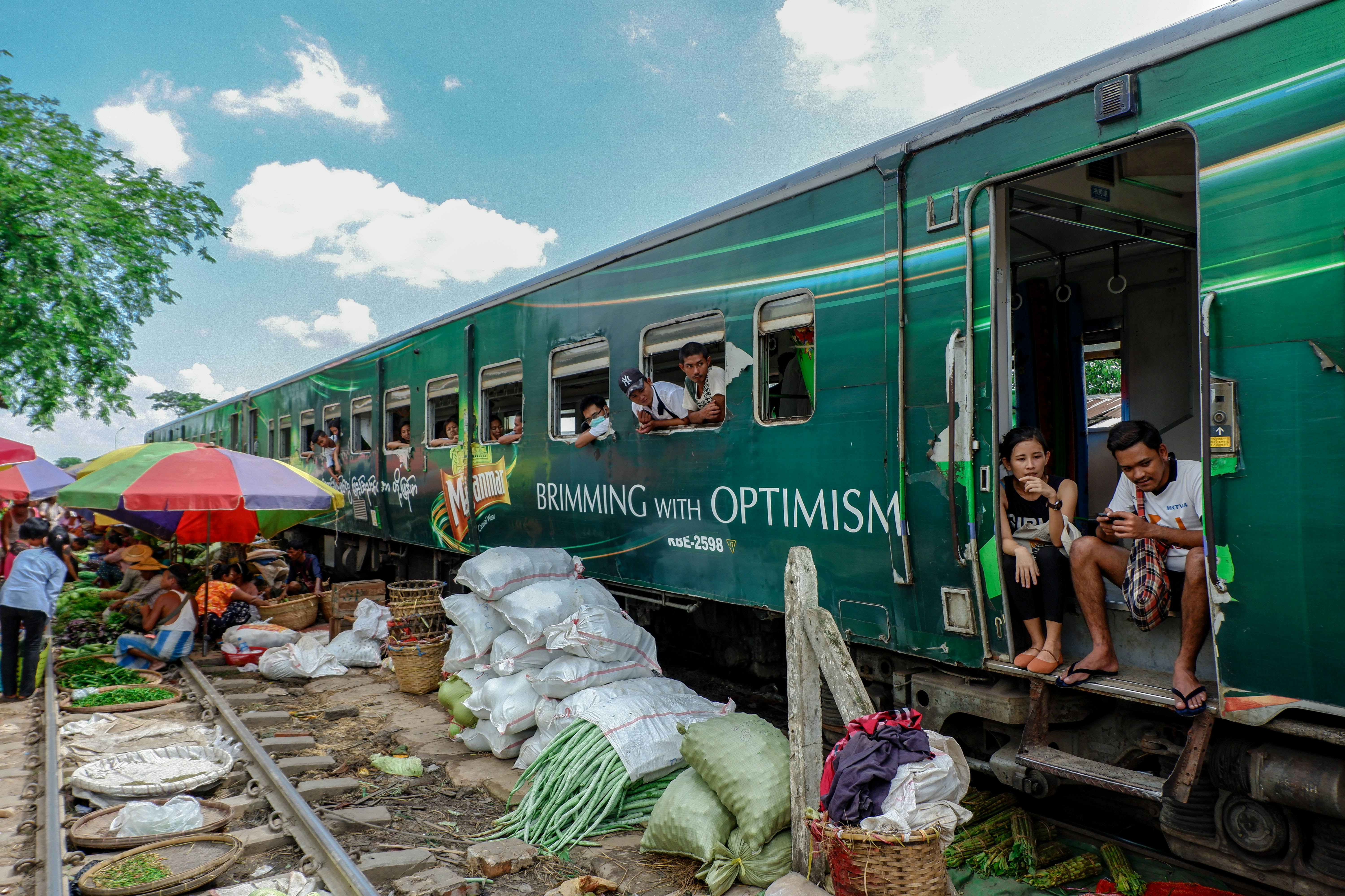 The green Yangon circle train is stationary on the tracks. Passengers are leaning out of the doors and windows to see the trackside stalls selling fresh produce.