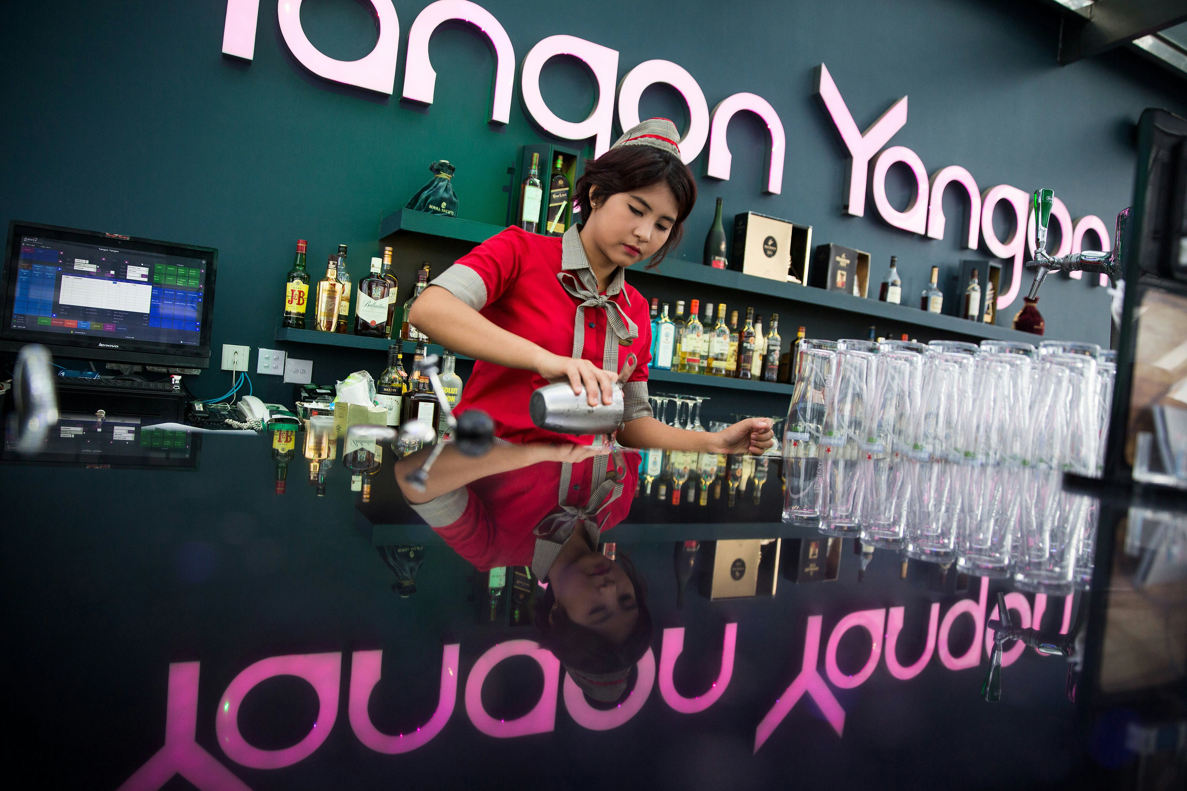 A bartender in uniform is preparing a cocktail in a fancy bar; a large sign on the wall behind her says 'Yangon Yangon'
