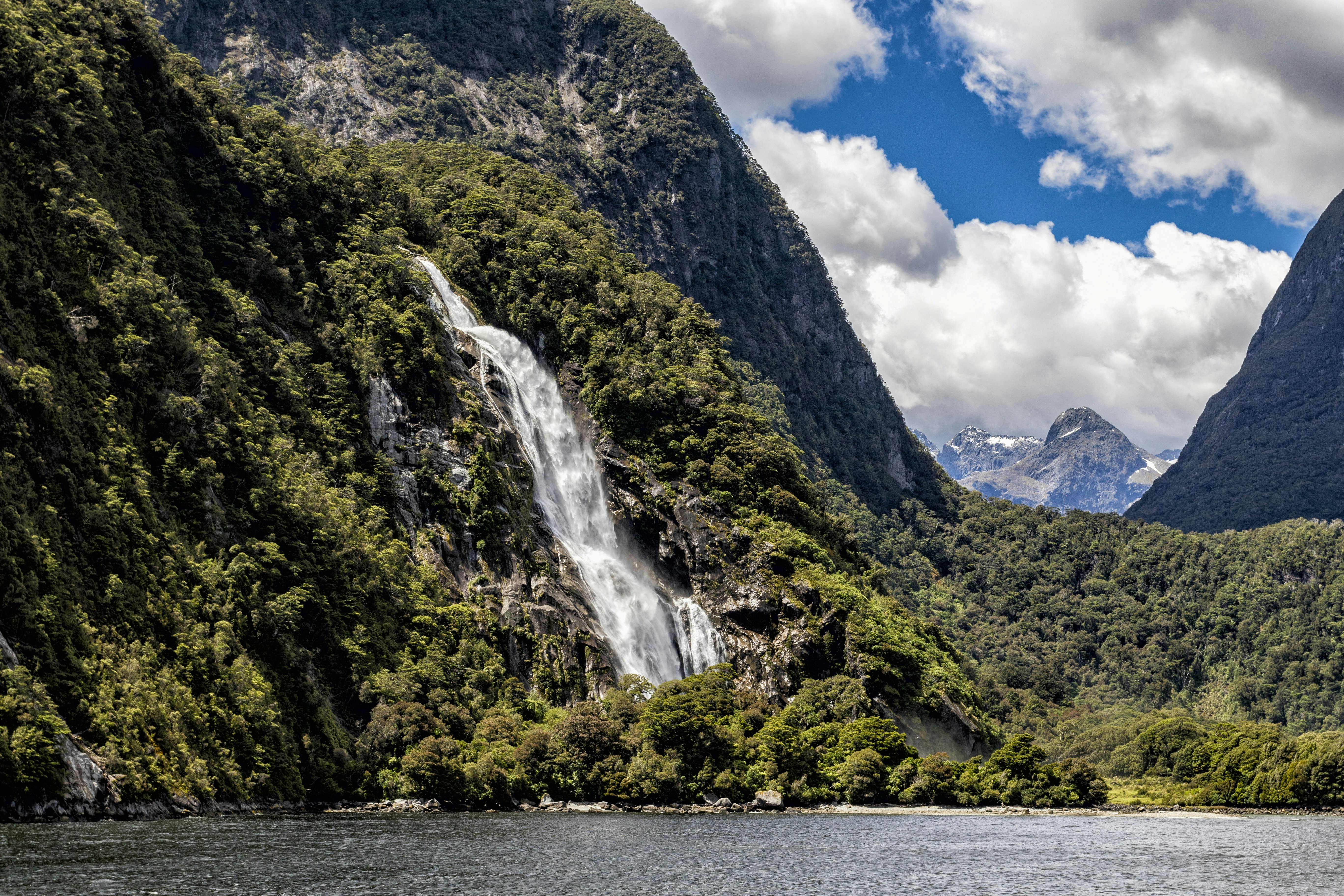 A bright white waterfall cascades down a deep green hillside in Fiordland National Park, South Island, New Zealand. In a dip between the steep hillsides, you can see blue snow-capped peaks in the distance. In the immediate foreground is water so blue that it's almost slate grey