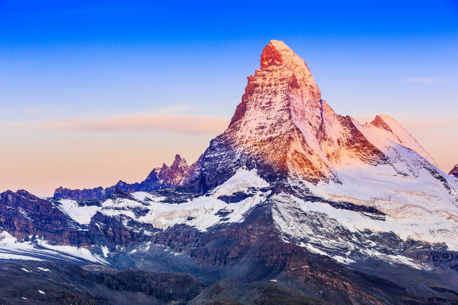 Looking much like a steeper-sided Egyptian pyramid, the chiseled flanks of the Matterhorn rise sharply into a blue sky; the who mountain and its snow-covered lower flanks glows warmly in the setting sun