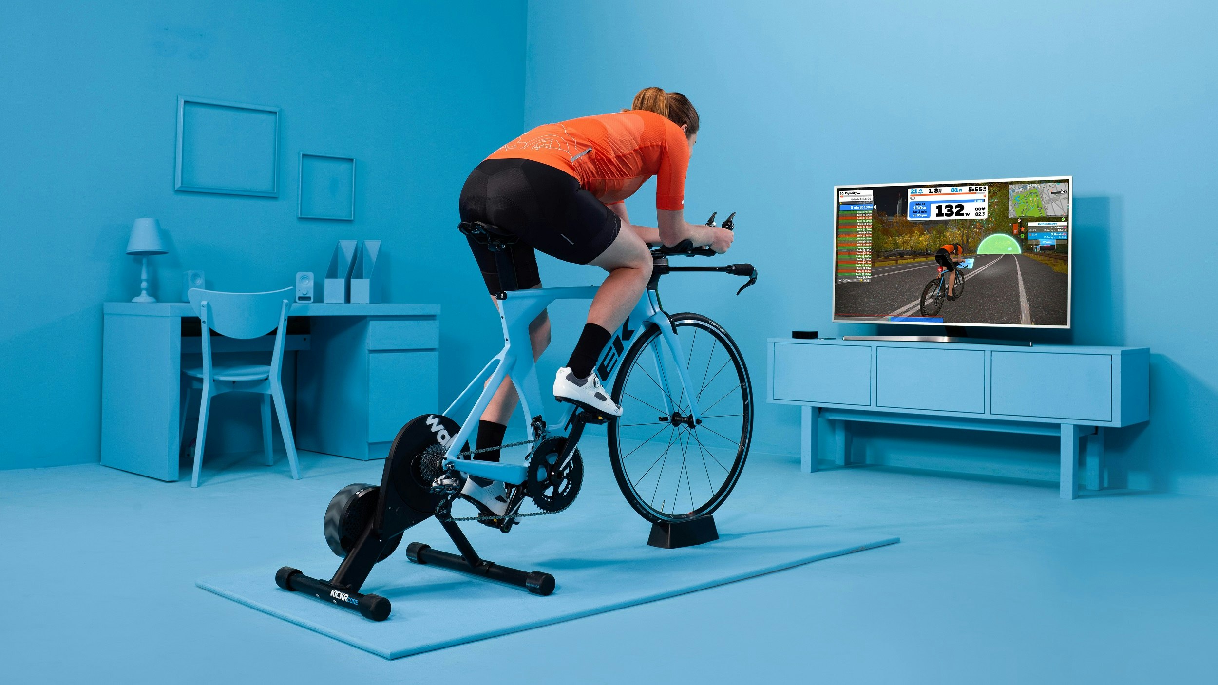 A woman rides a time-trial bike on a turbo trainer in front of a TV; on the TV is her avatar in the Zwift cycling race.