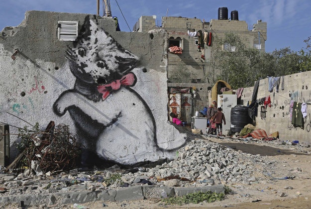 A Palestinian woman works with her children near a mural of a playful-looking kitten, presumably painted by British street graffiti artist Banksy.